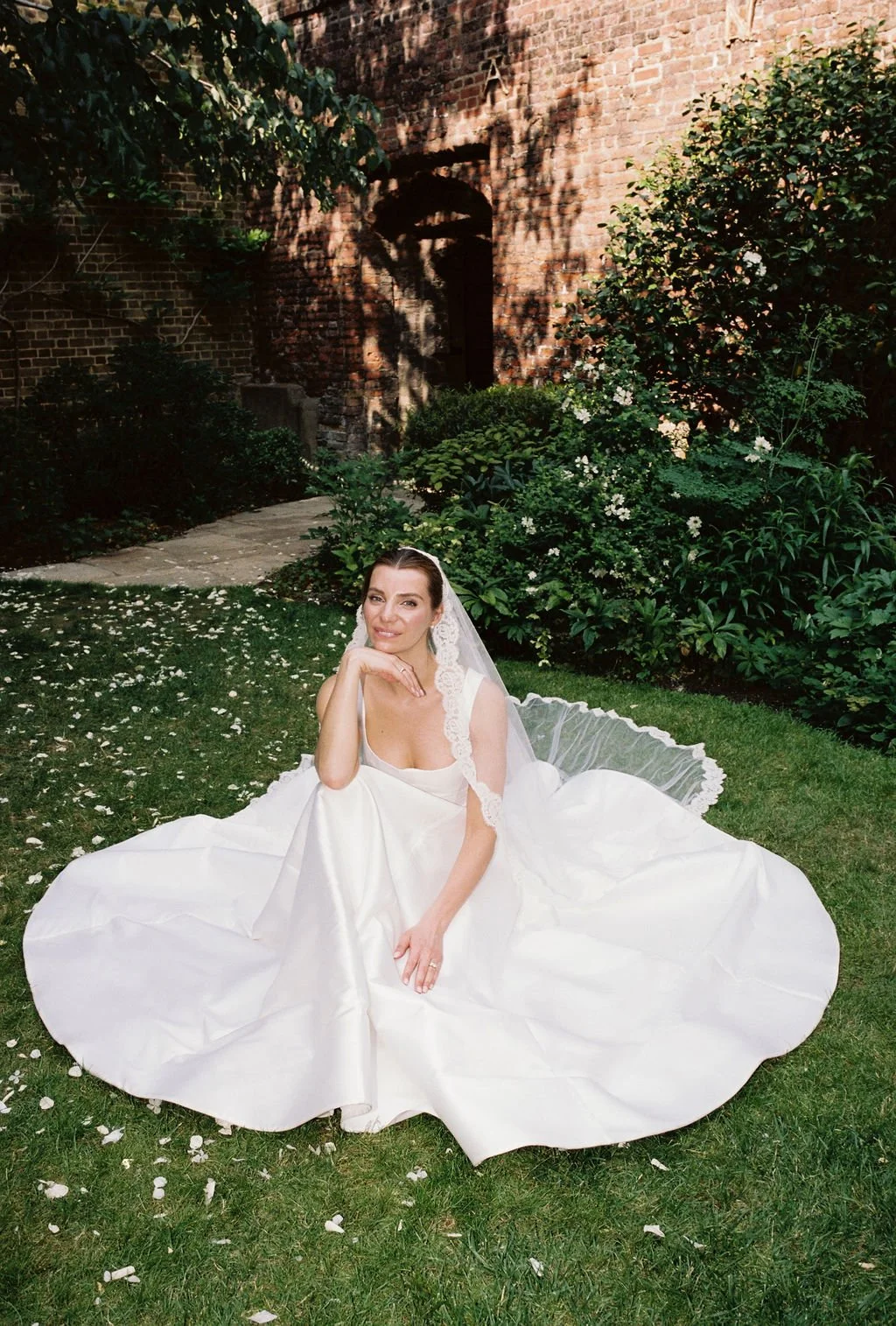 A woman in a wedding dress sitting on grass in a garden with green bushes and a brick wall. She has a veil and is looking at the camera with a smile.
