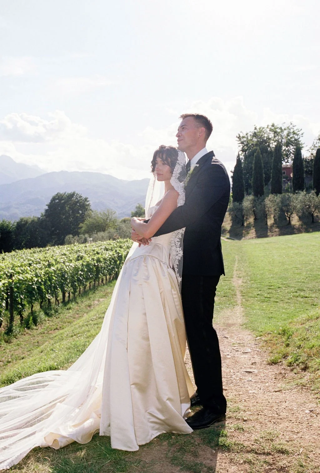 A bride and groom standing in a vineyard on their wedding day, with mountains and trees in the background.