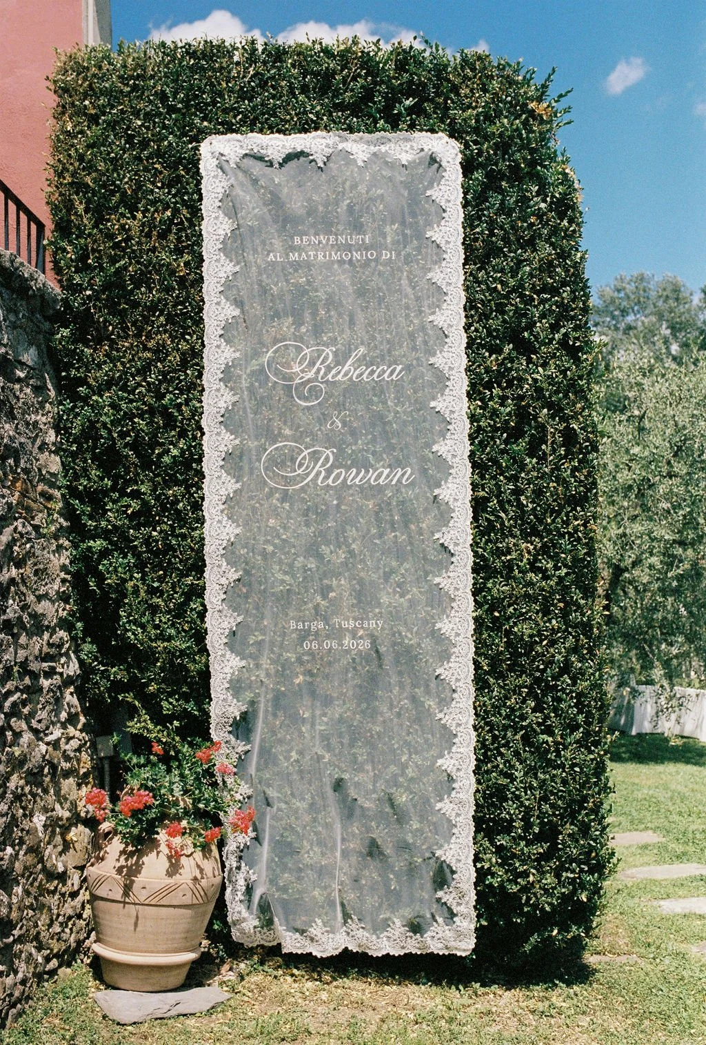 Wedding welcome sign with lace border, displaying the names Rebecca and Rowan, in Tuscany on June 6, 2026, outdoors among greenery.