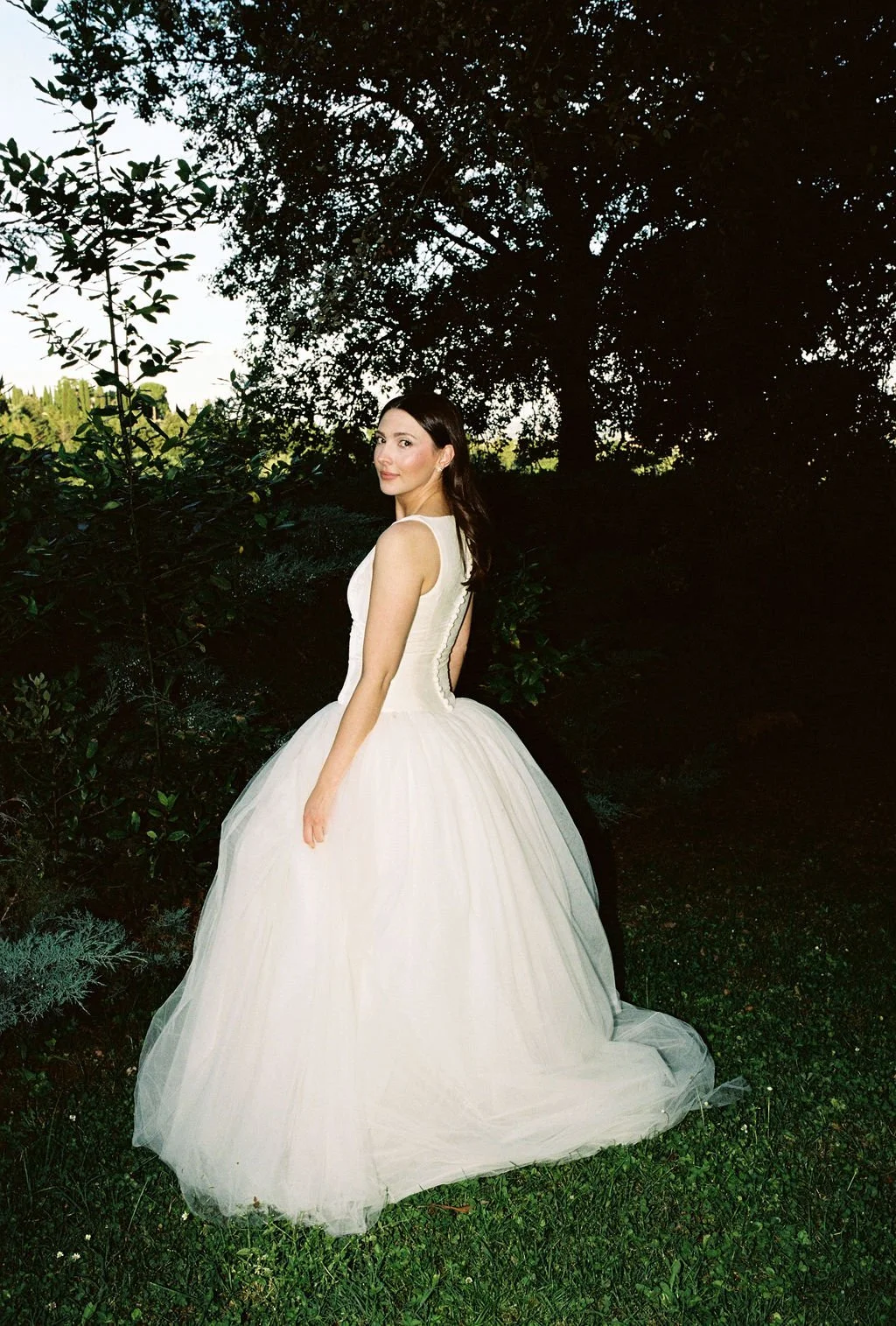 A woman in a white wedding gown standing outdoors in a lush, green setting with trees and bushes, under a bright sky.