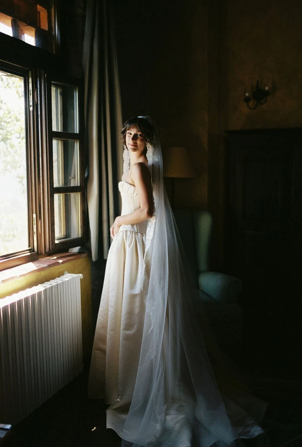 A bride standing indoors near a window, wearing a white wedding dress and a veil, looking towards the camera with natural light illuminating her face.