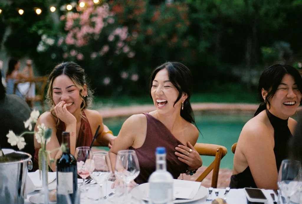Three women laughing and having fun at a dinner party outdoors during evening, with string lights and pink flowers in the background.