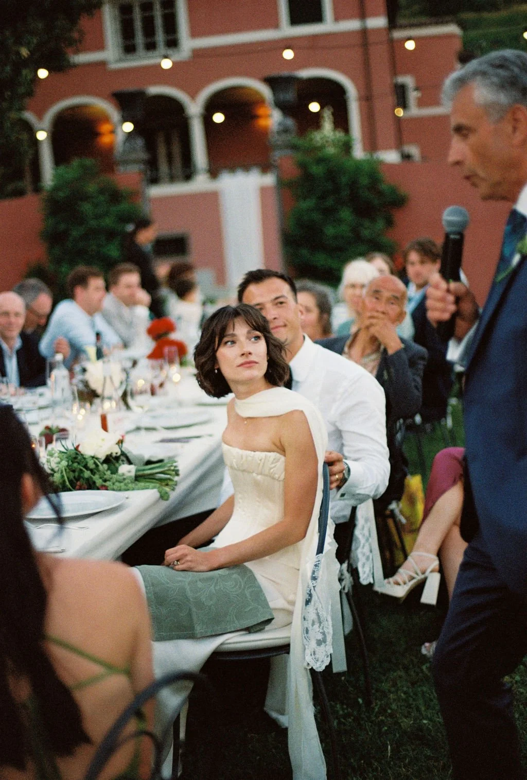 A woman in a white strapless dress and a man in a white shirt sitting at head of a long dinner table during an outdoor wedding reception, listening to a man in a blue suit giving a speech with a microphone, as other guests watch.