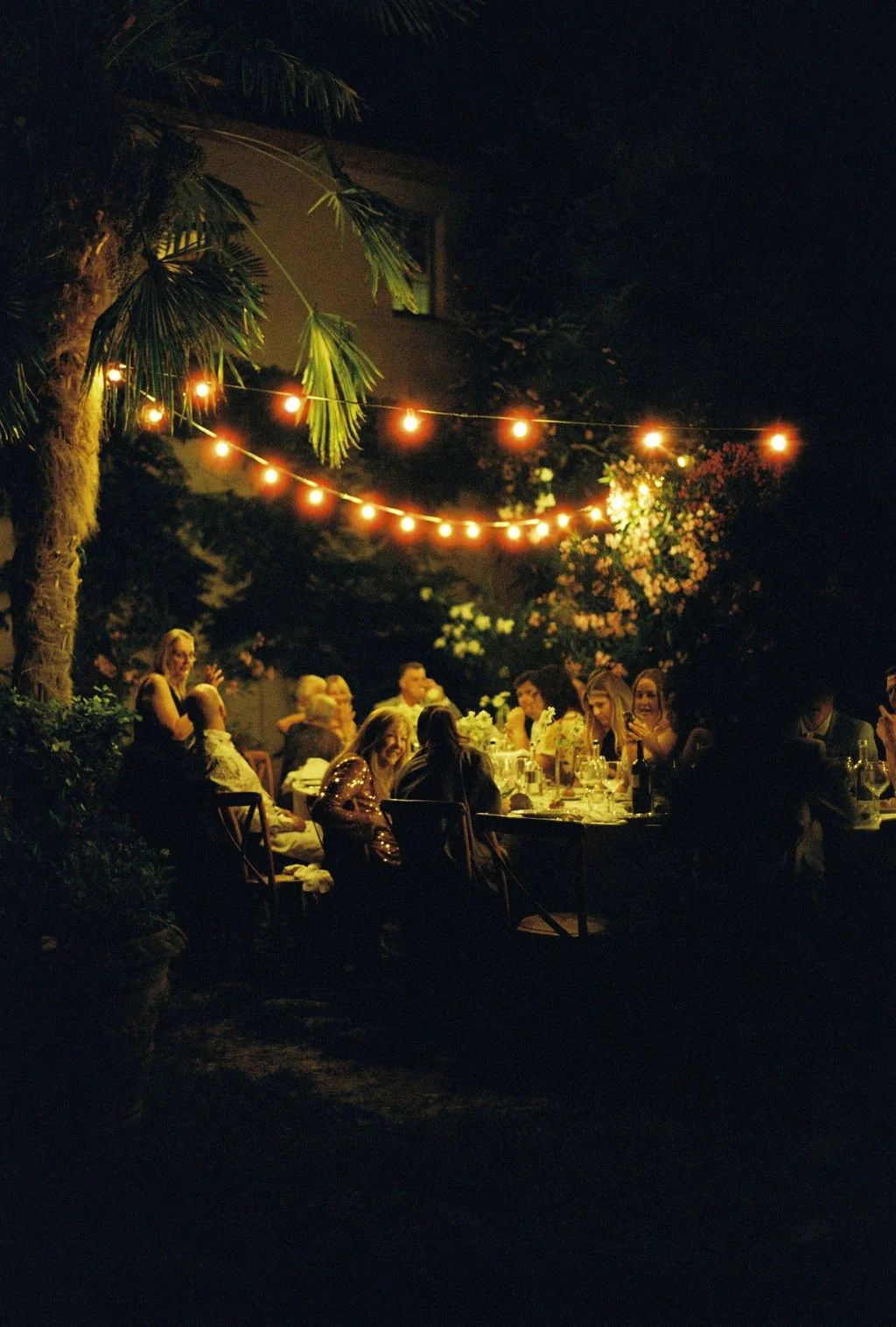 People gather at a nighttime outdoor dinner party under string lights, surrounded by trees and flowers.