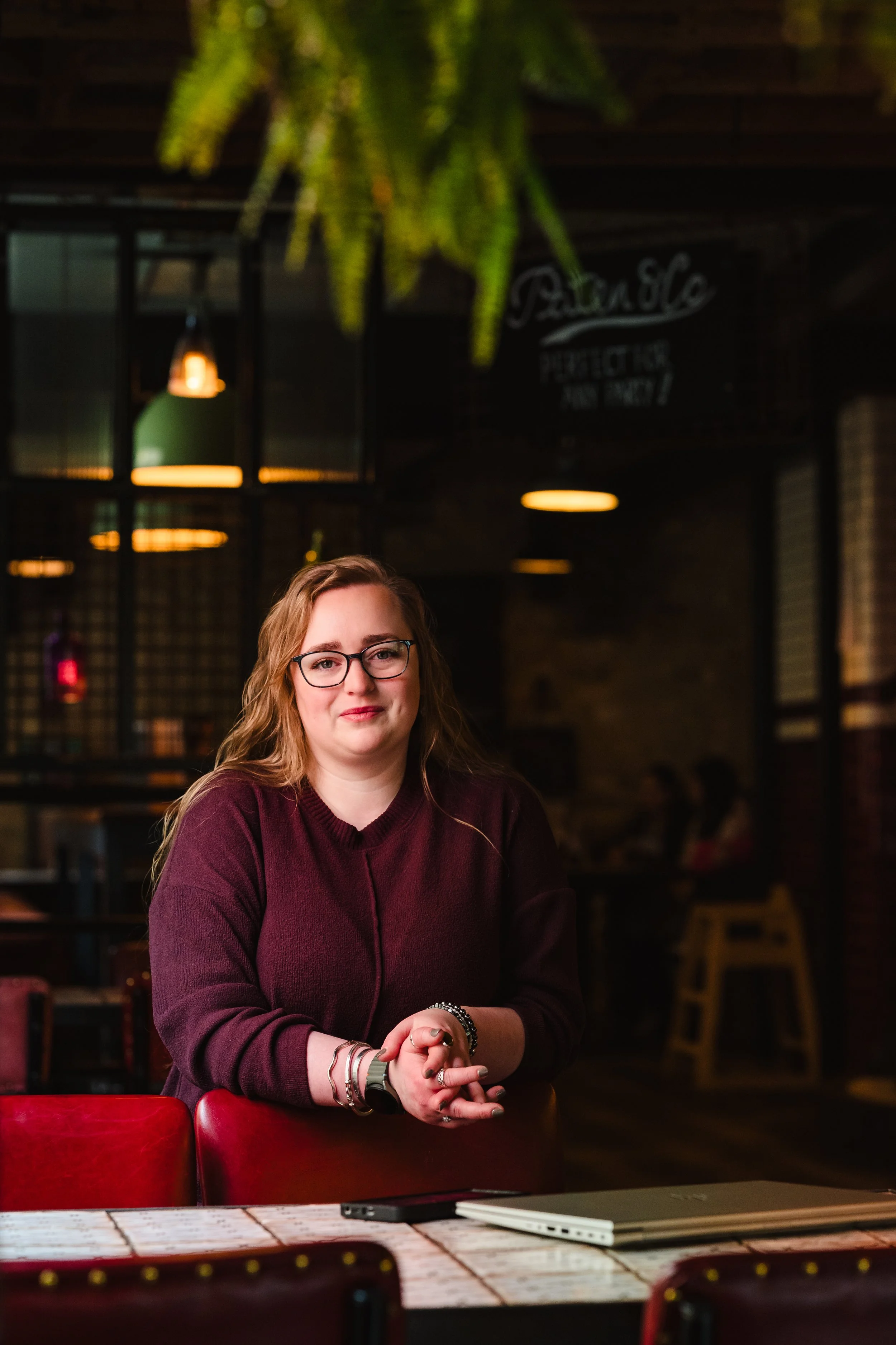 A woman with glasses and long wavy hair sitting at a table in a restaurant or cafe, wearing a maroon sweater and jewelry, with a green plant hanging above her and warm ambient lighting in the background.