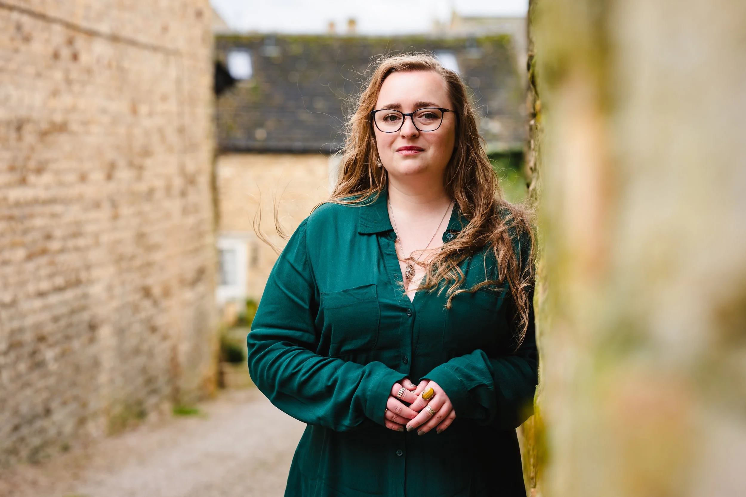 A woman with long wavy hair, wearing glasses and a dark green button-up shirt, standing outdoors between brick and moss-covered walls, looking at the camera with a neutral expression.