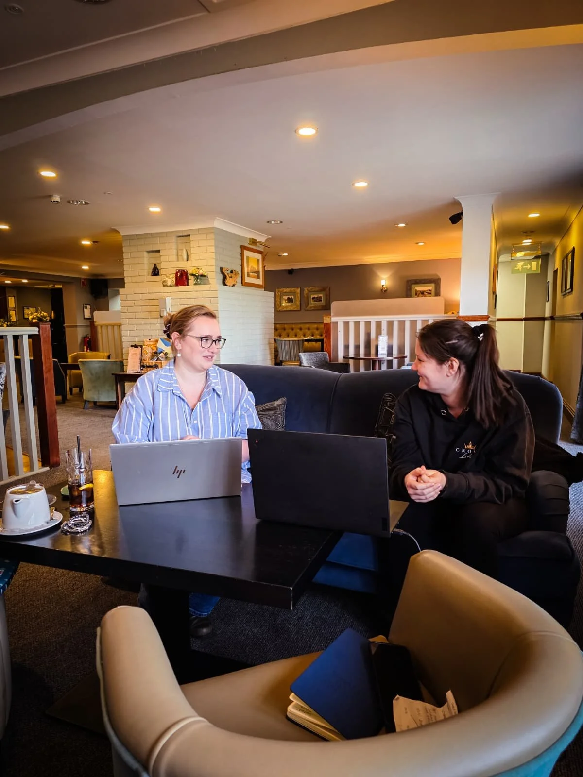 Two women having a conversation while sitting at a table with laptops in a cozy, well-lit restaurant or cafe. One woman is wearing glasses and a striped shirt, the other woman is in a black hoodie.
