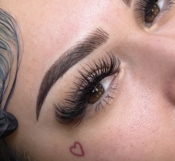 Close-up of a person's eye with long eyelashes, arched eyebrows, and a small heart tattoo below the eye.