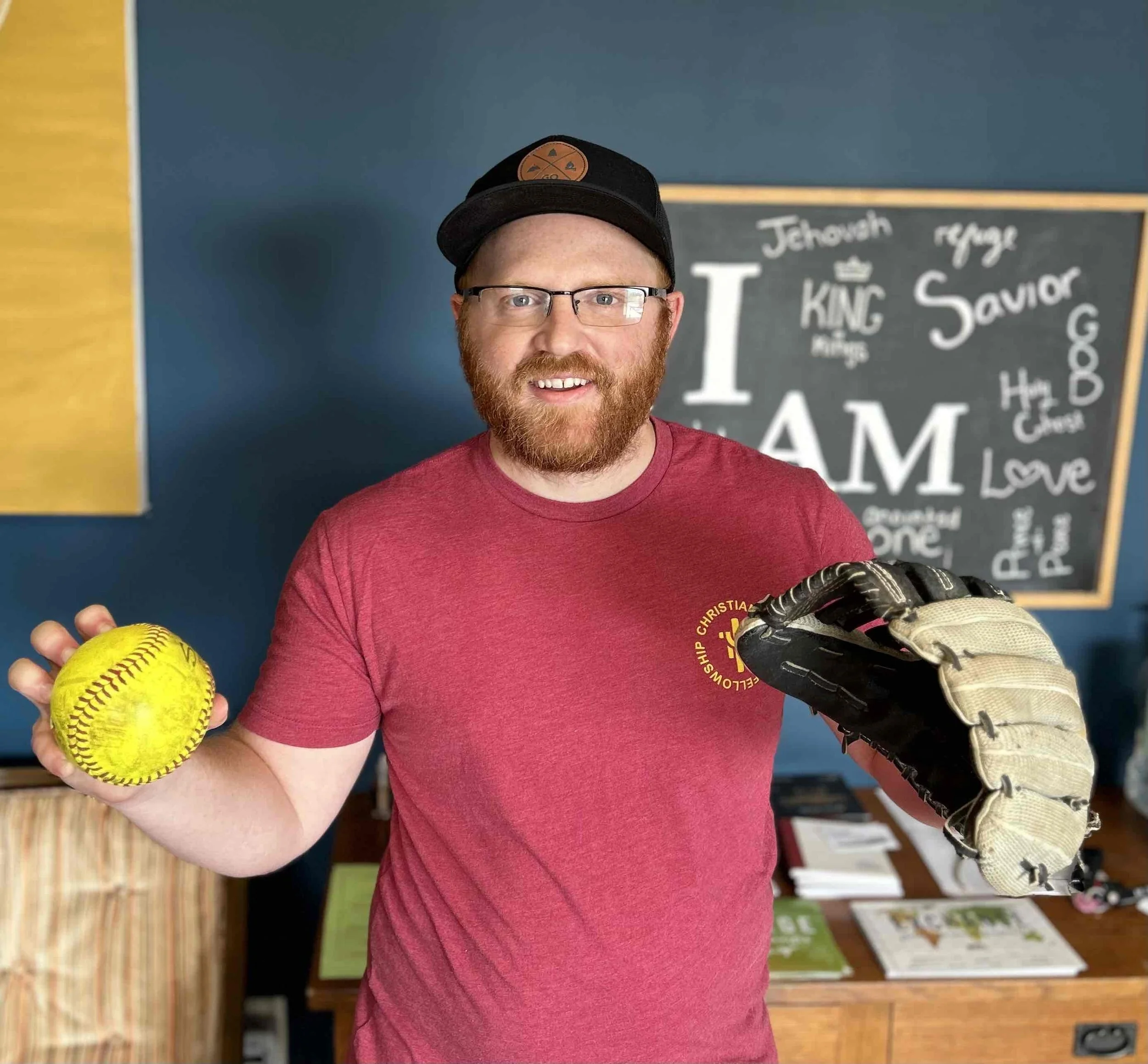 A man with glasses and a red beard holding a yellow softball in his right hand and a baseball glove in his left hand, standing in a room with blue walls and a blackboard in the background.