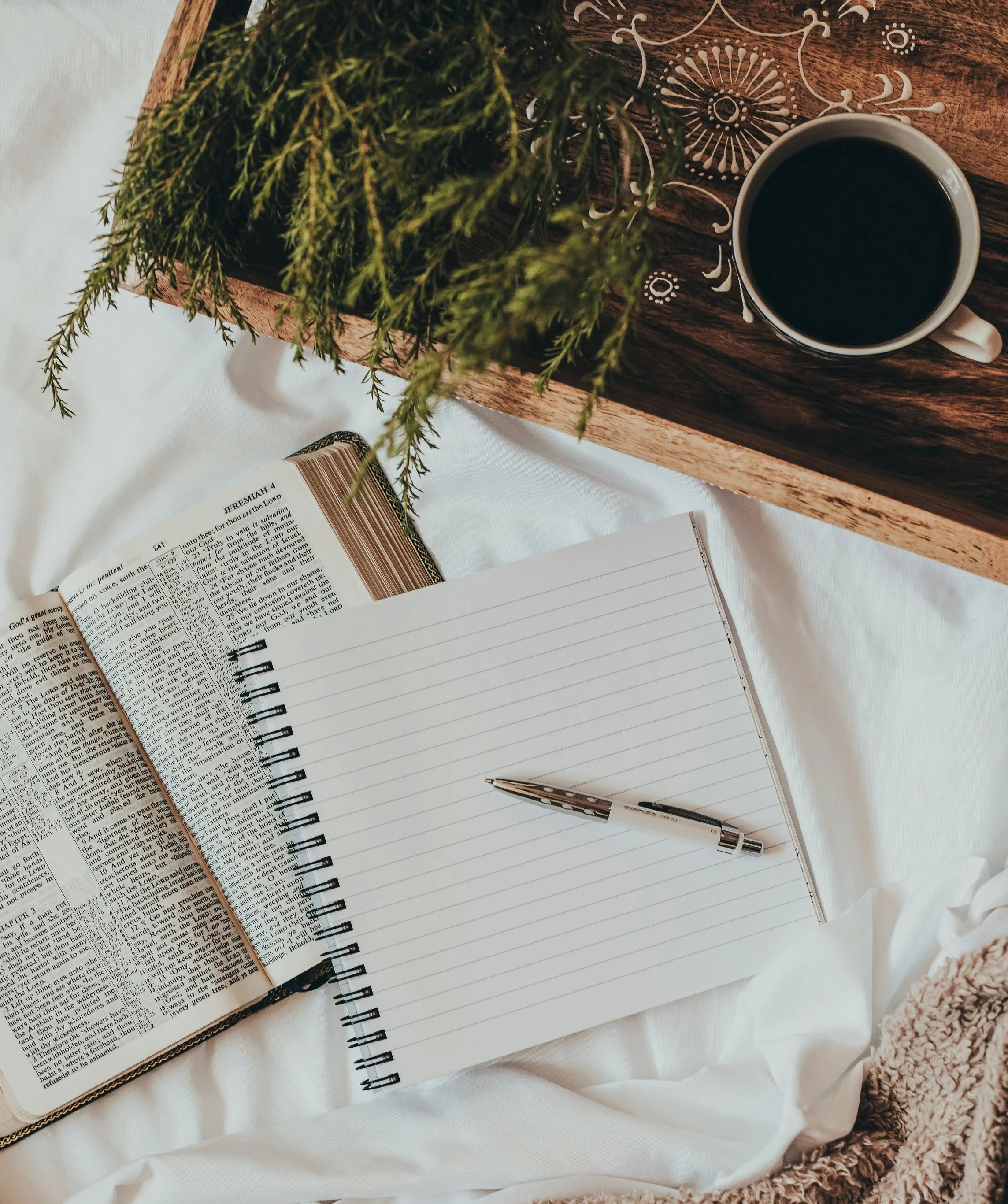 Open Bible, blank notebook with pen, cup of black coffee, and potted plant on a wooden tray, all arranged on a white fabric surface.
