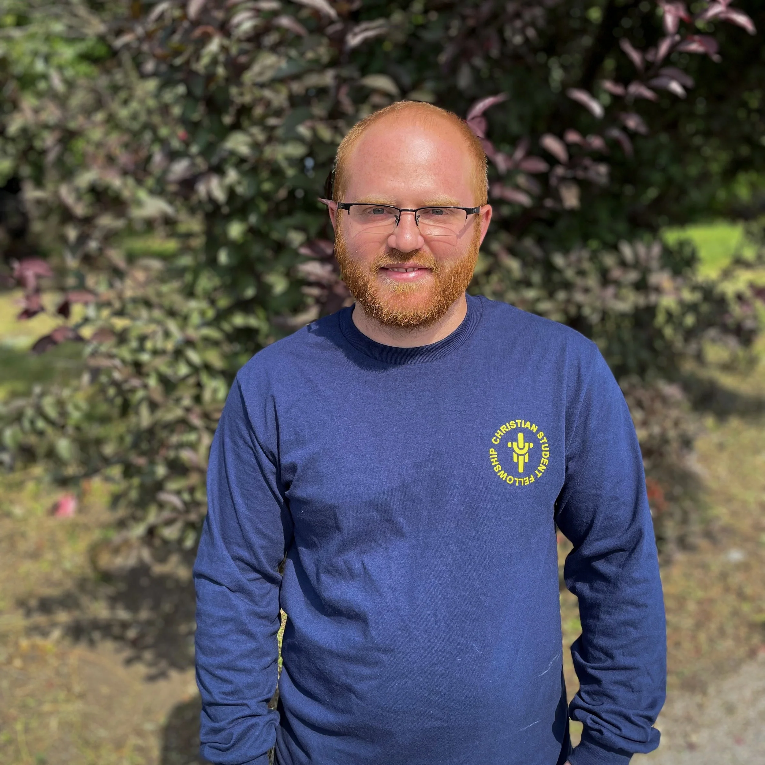 A man with glasses, red hair, and a beard standing outdoors in front of a bush, wearing a navy blue long-sleeve shirt with a yellow Christian symbol and text on the chest.