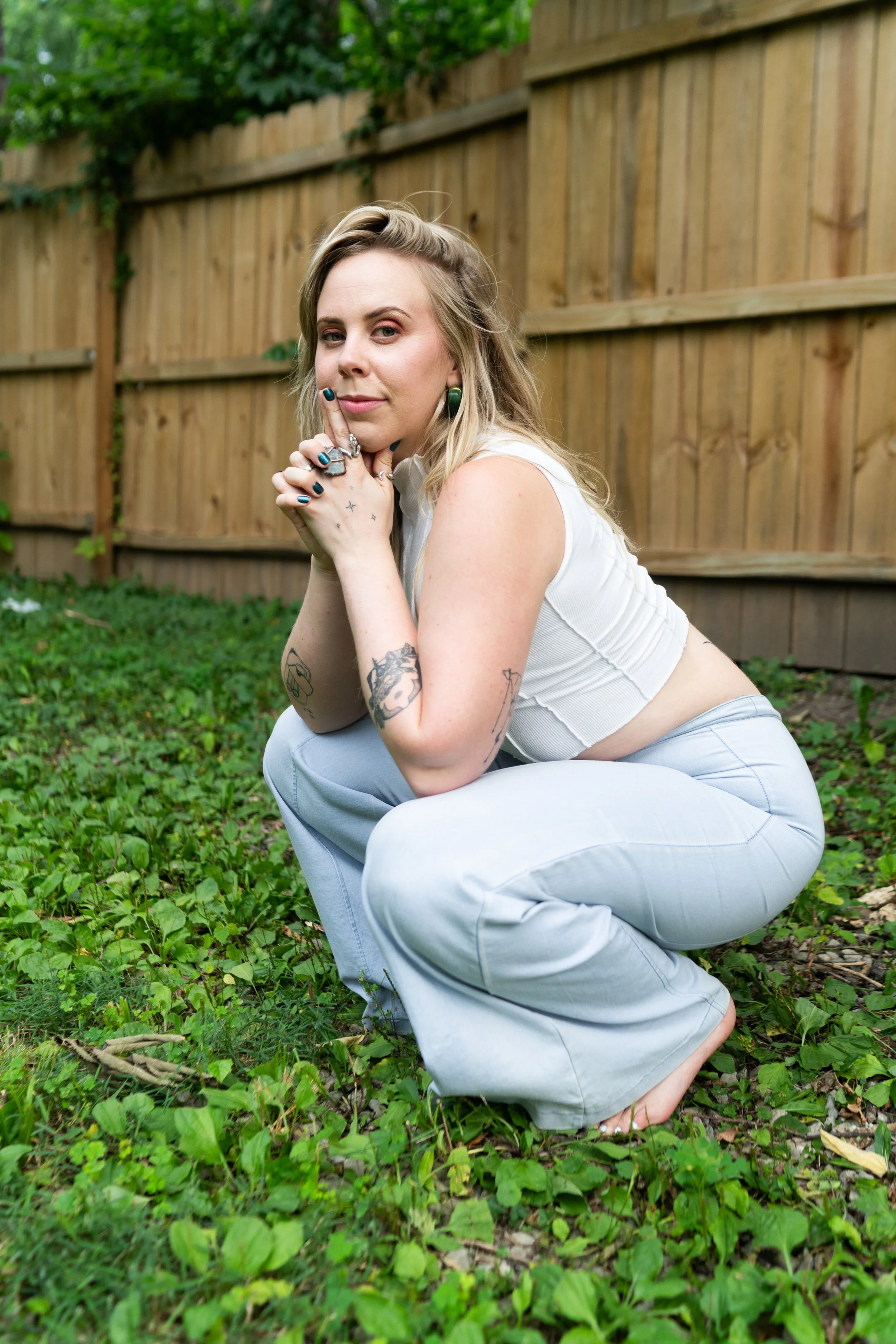 A portrait of Erin wearing  a white ribbed tank top, chunky green earrings, and wide-legged light blue jeans.