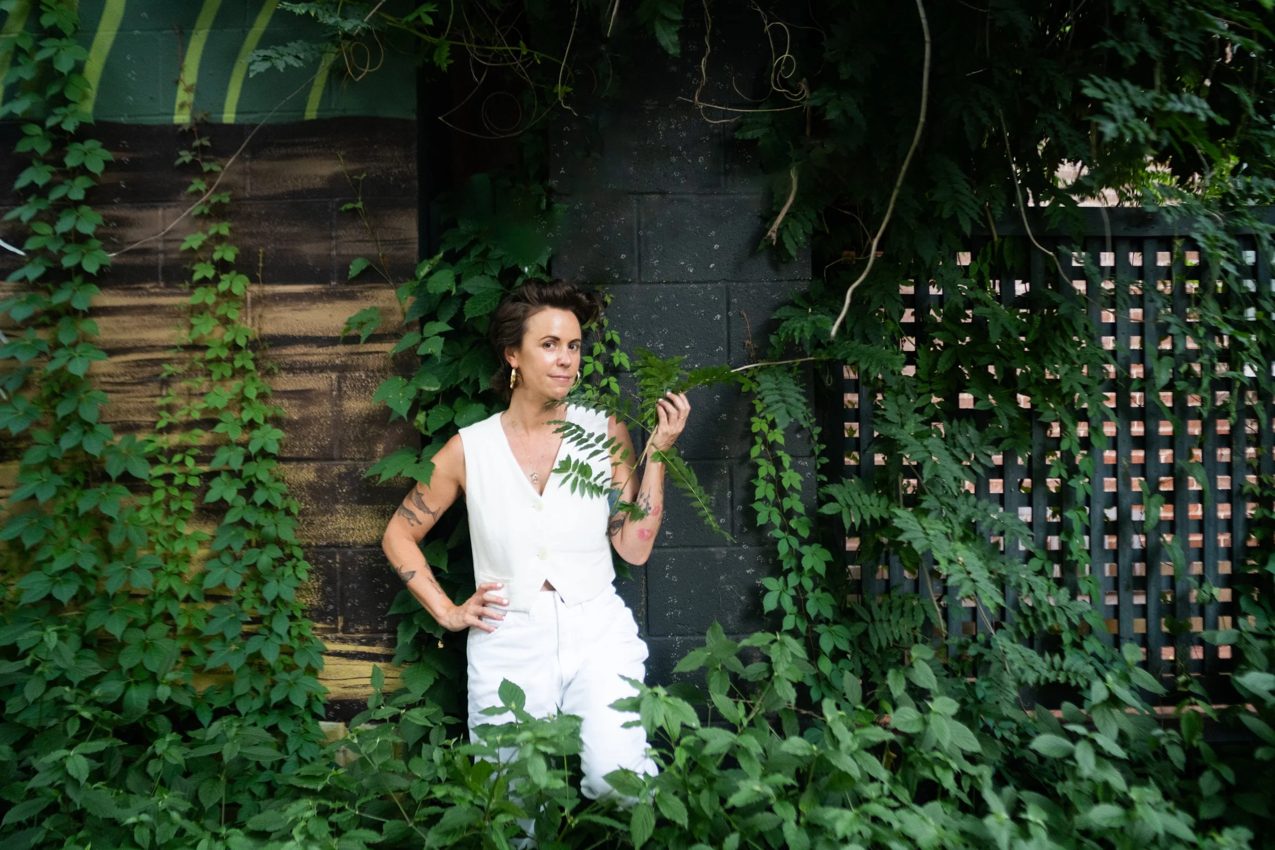 A portrait of Jardana in a white vest, white jeans, and gold hoop earings leaning against painted black brick and surrounded by lush green.