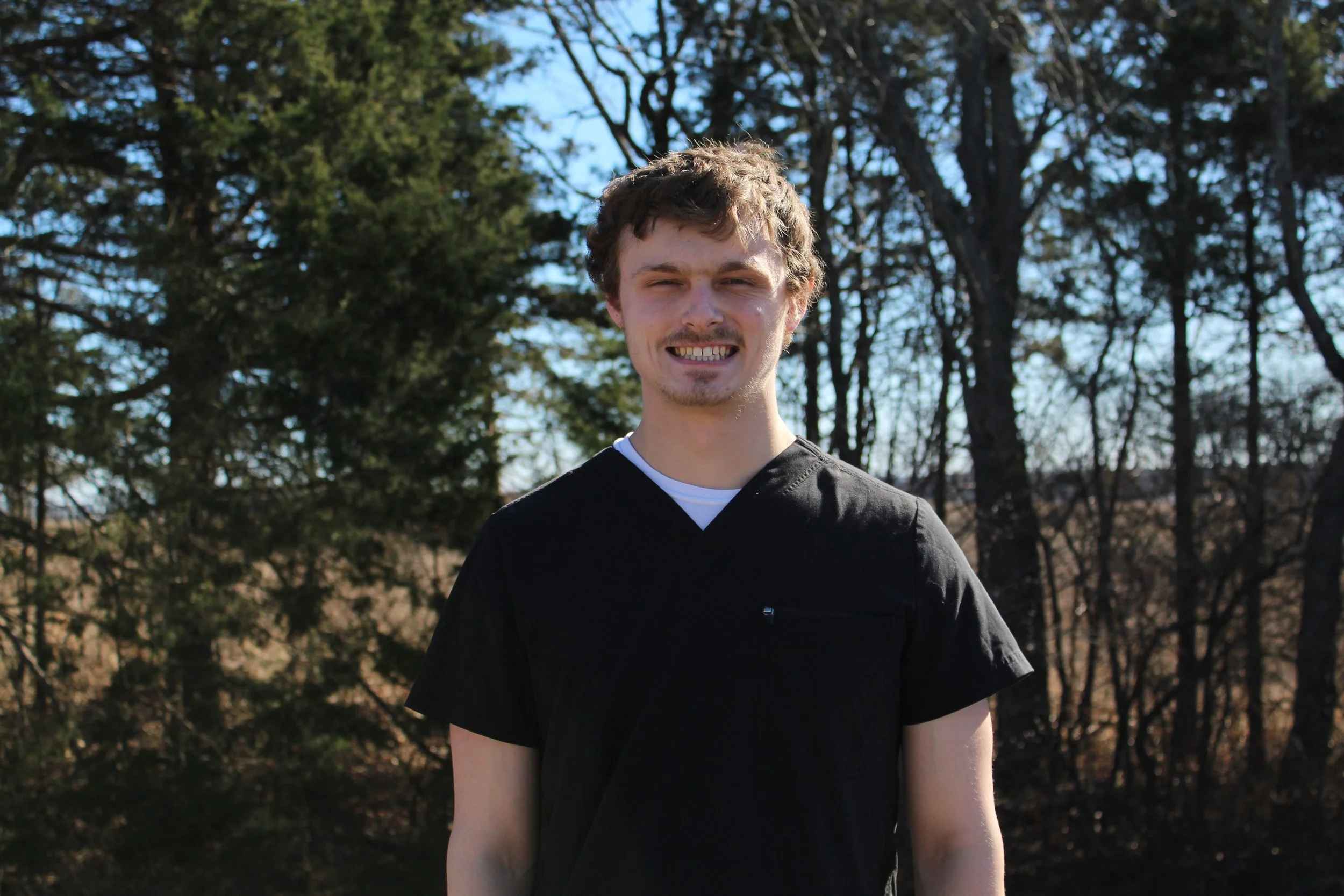 A young man smiling outdoors with trees and a clear blue sky in the background.