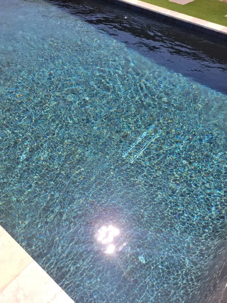 Clear swimming pool water with sunlight reflecting, bordered by tiles and a grassy area.
