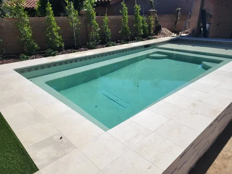 Backyard swimming pool with stone tile surround and greenery.