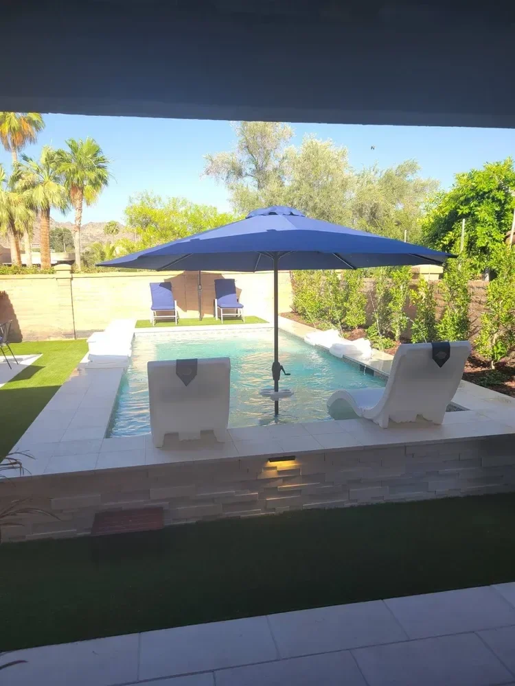 Backyard pool area with a rectangular swimming pool, blue lounge chairs, a large blue umbrella, and surrounding greenery including palm trees and shrubs.