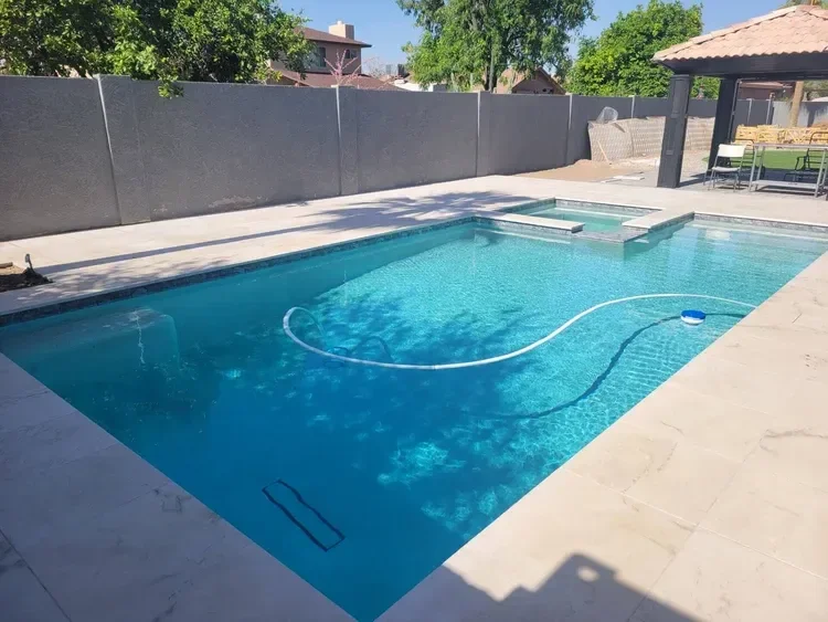 Residential outdoor swimming pool with clear blue water, surrounded by a light-colored stone deck, and partially shaded by nearby trees and a small structure with a tiled roof. Large wall and greenery in the background.