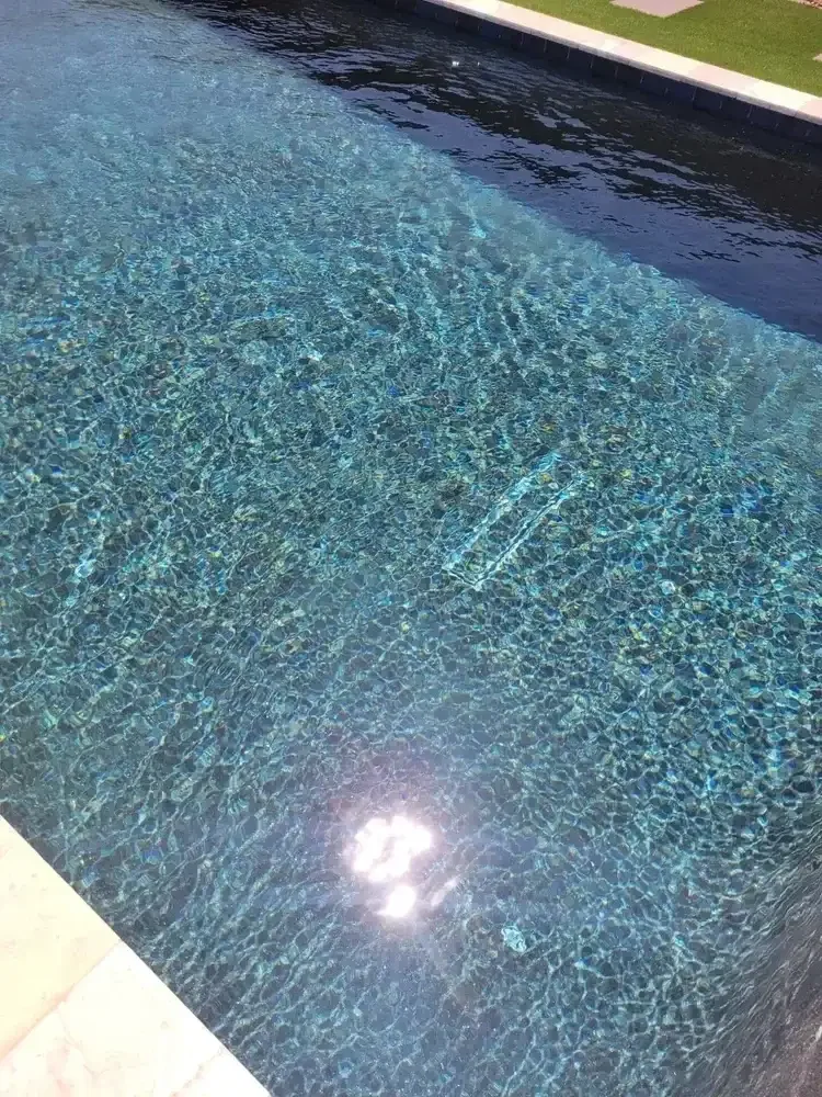 Clear swimming pool water with sunlight reflecting, bordered by tiles and a grassy area.