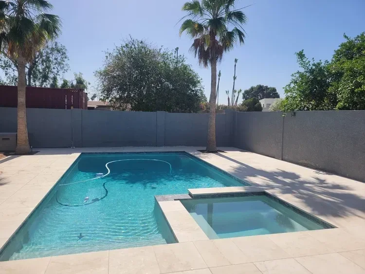 Backyard with a swimming pool and hot tub, surrounded by palm trees and gray fence.