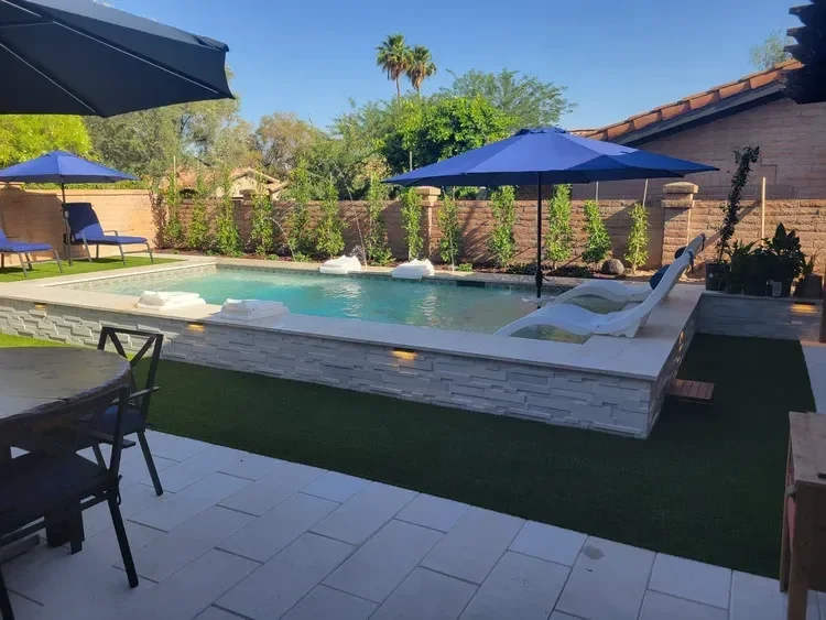 Backyard with a small rectangular pool, white lounge chairs, blue umbrellas, patio table and chairs, green artificial grass, surrounded by brick walls and trees.