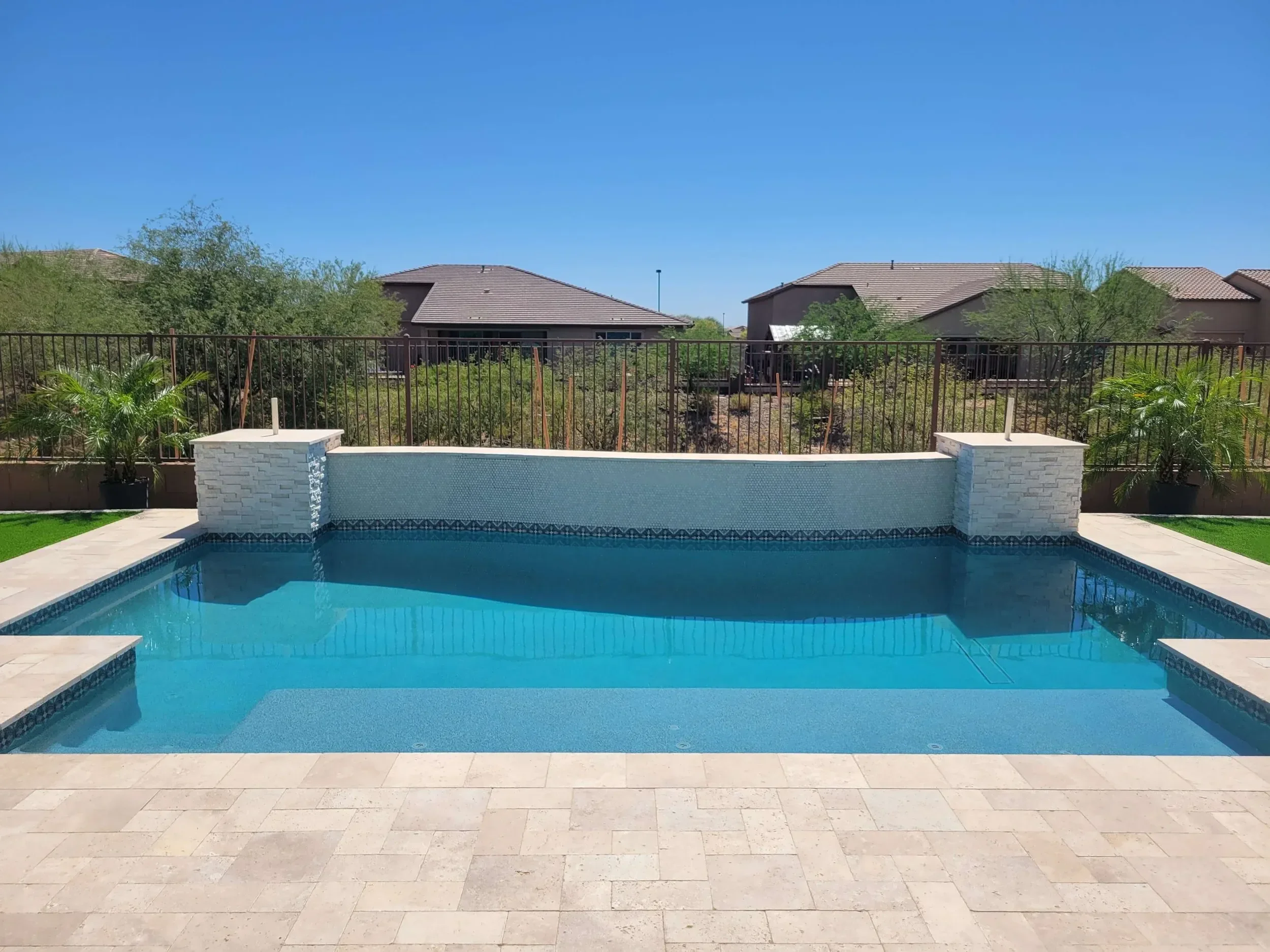 Outdoor swimming pool with clear blue water, surrounded by a tiled patio and decorative stone walls. In the background, there is a metal fence with potted plants, and beyond the fence, residential houses and shrubs are visible under a clear blue sky.