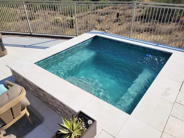 A small outdoor pool with clear blue water, surrounded by a stone and tile deck. There's a patio chair with cushions nearby and a potted plant in the foreground. A metal fence encloses the area, and the background shows a dry, desert landscape.