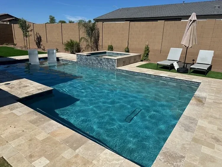Backyard swimming pool with lounge chairs, umbrella, and a stone patio under clear blue sky.