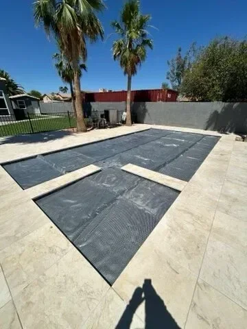 Swimming pool covered with a pool cover, surrounded by a stone patio, with palm trees in the background.