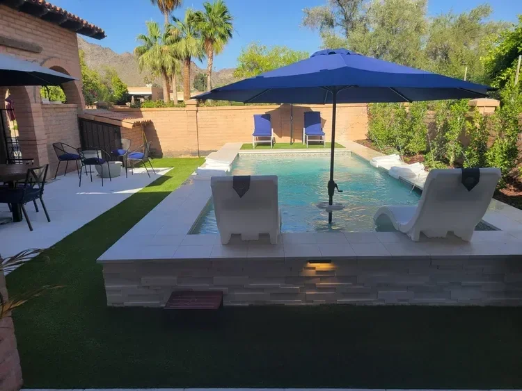 Backyard pool area with white loungers, blue umbrellas, and patio furniture. Surrounded by greenery and a mountain view.