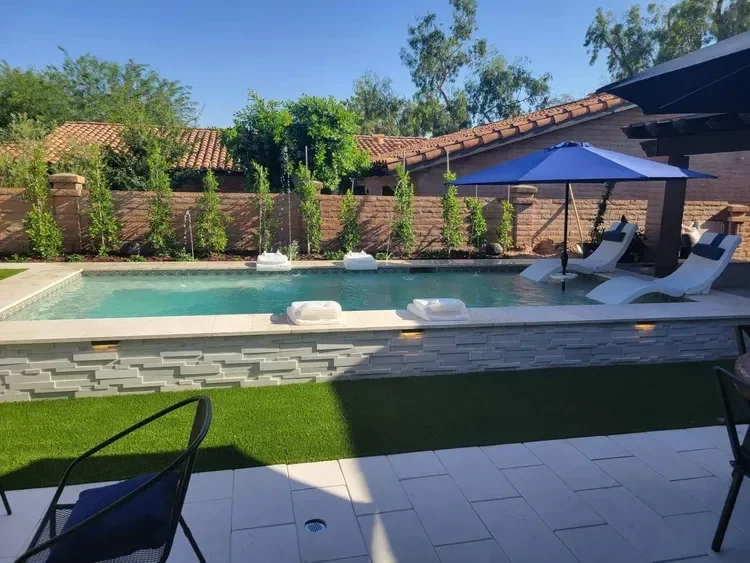 Backyard swimming pool with lounge chairs, blue umbrella, and greenery against a brick wall.