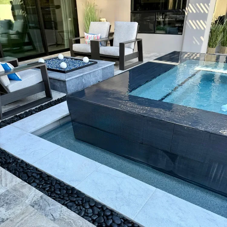 Modern outdoor patio with a small pool, black stone edging, gray armchairs, and a fire feature with decorative balls. Plants and potted grass complete the contemporary design.