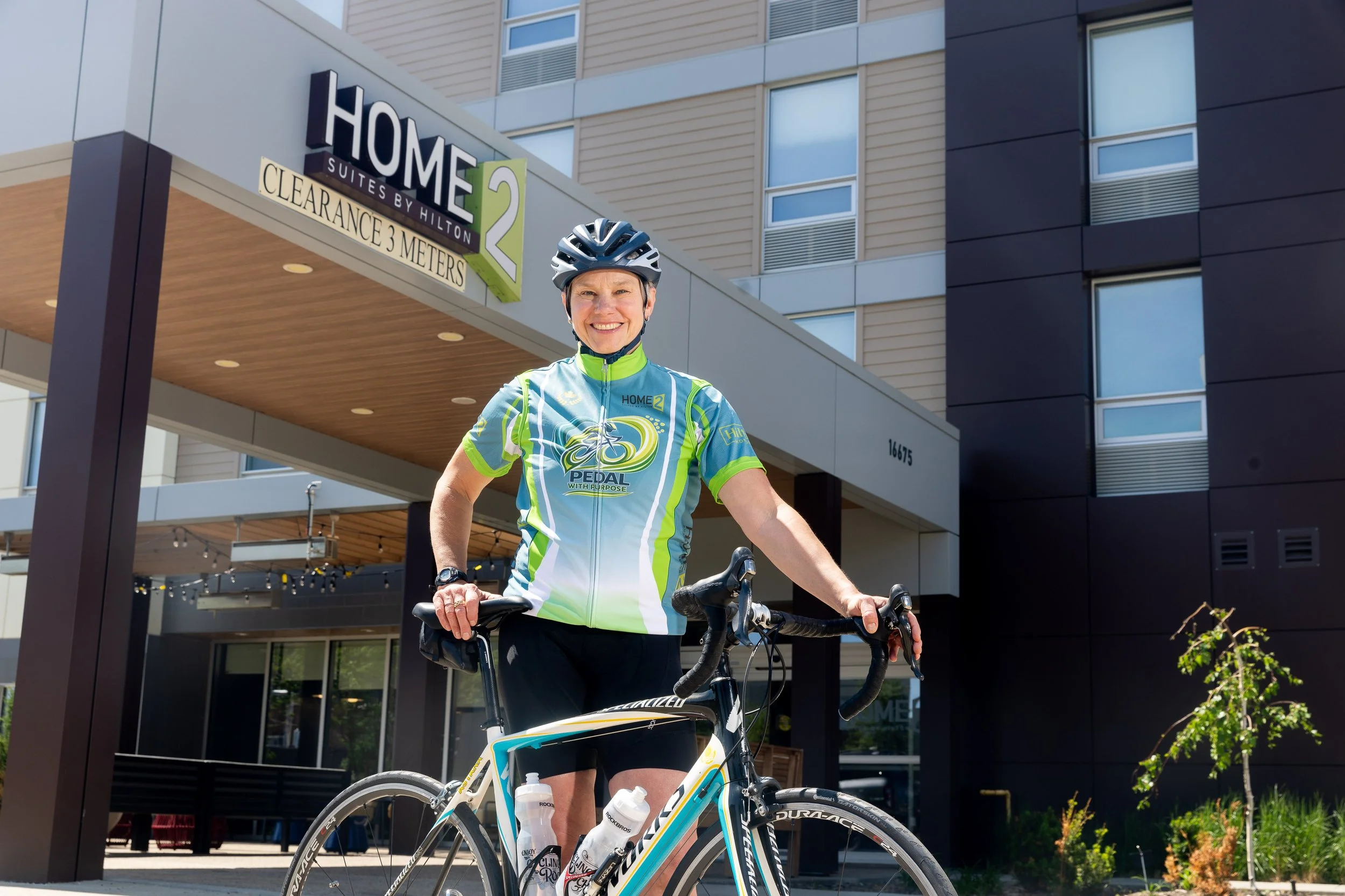 A smiling woman wearing a cycling helmet and colorful cycling jersey stands with her bicycle in front of a modern city building with large windows and a sign that reads 'Home 2 Suites by Hilton.'