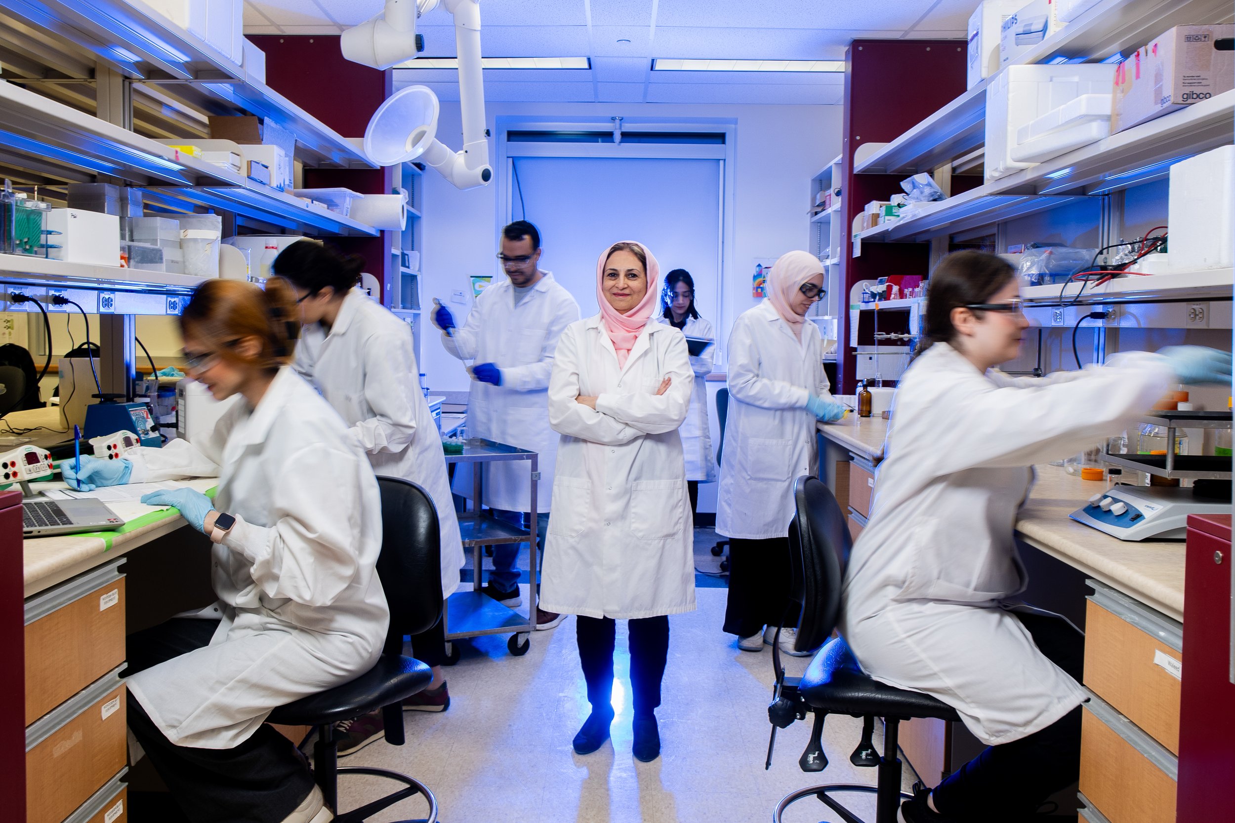 Scientists working in a laboratory with shelves of equipment and supplies. One woman stands with arms crossed and smiling at the camera, other scientists are engaged in work at the benches.