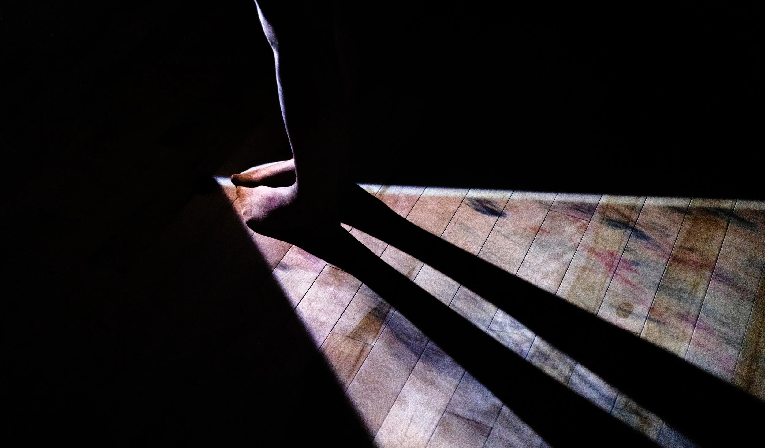 Silhouette of a person's legs and feet cast in shadow on a wooden floor in a dim room with a beam of light illuminating part of the floor.