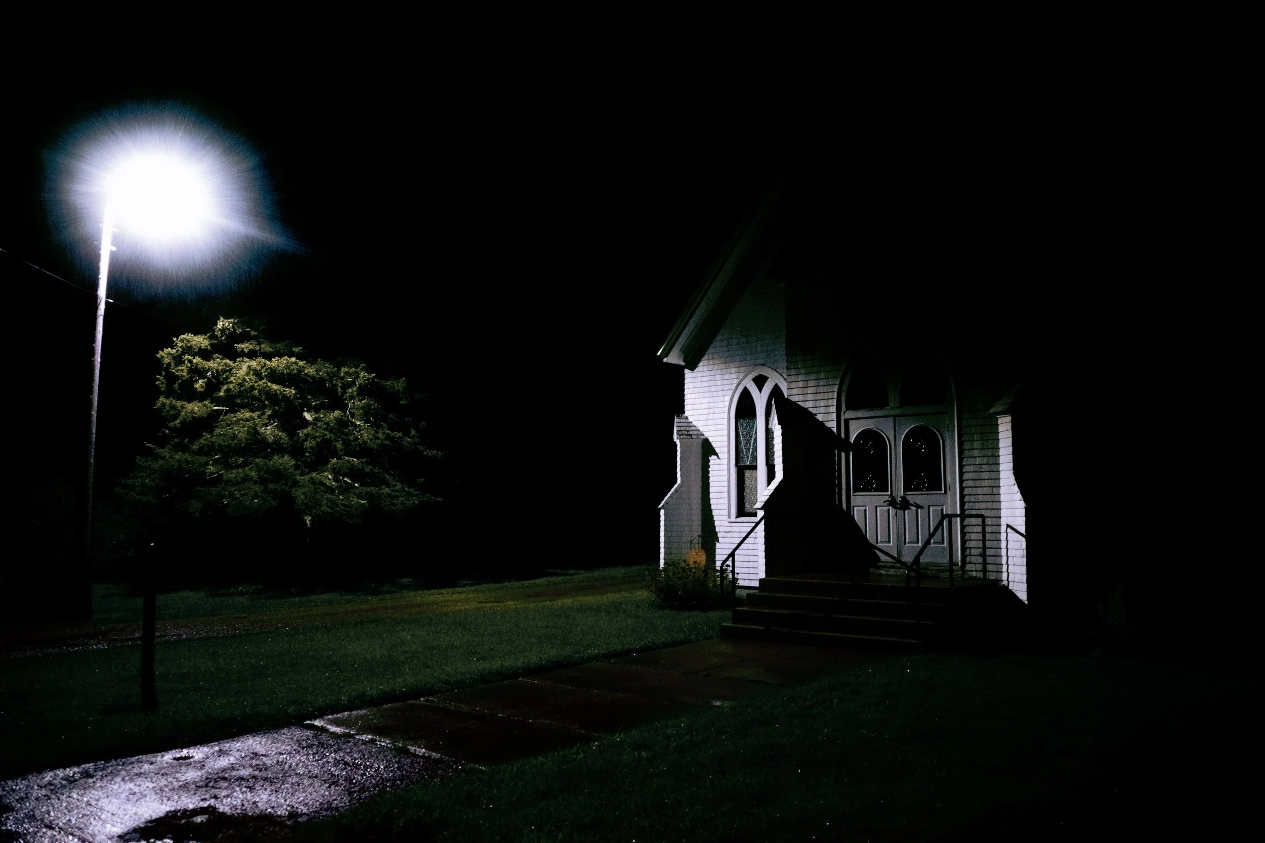 Nighttime scene of a small white church illuminated by streetlight with steps leading up to double doors, a tree nearby, and wet pavement reflecting light.