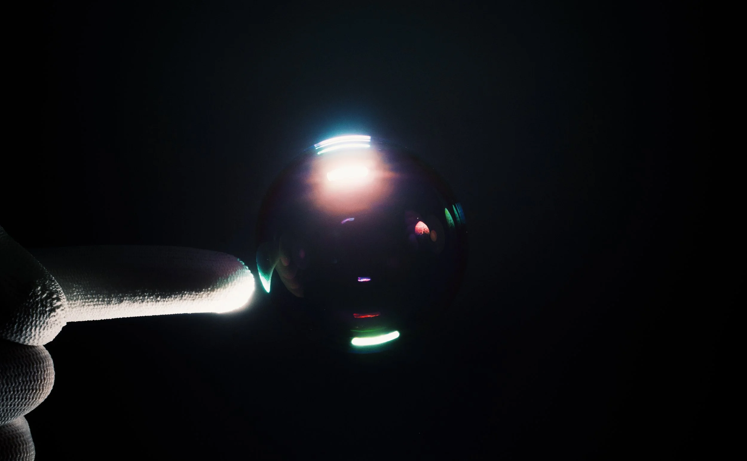 A gloved hand holding a reflective glass sphere in darkness, with light reflections creating colorful rings on the surface.