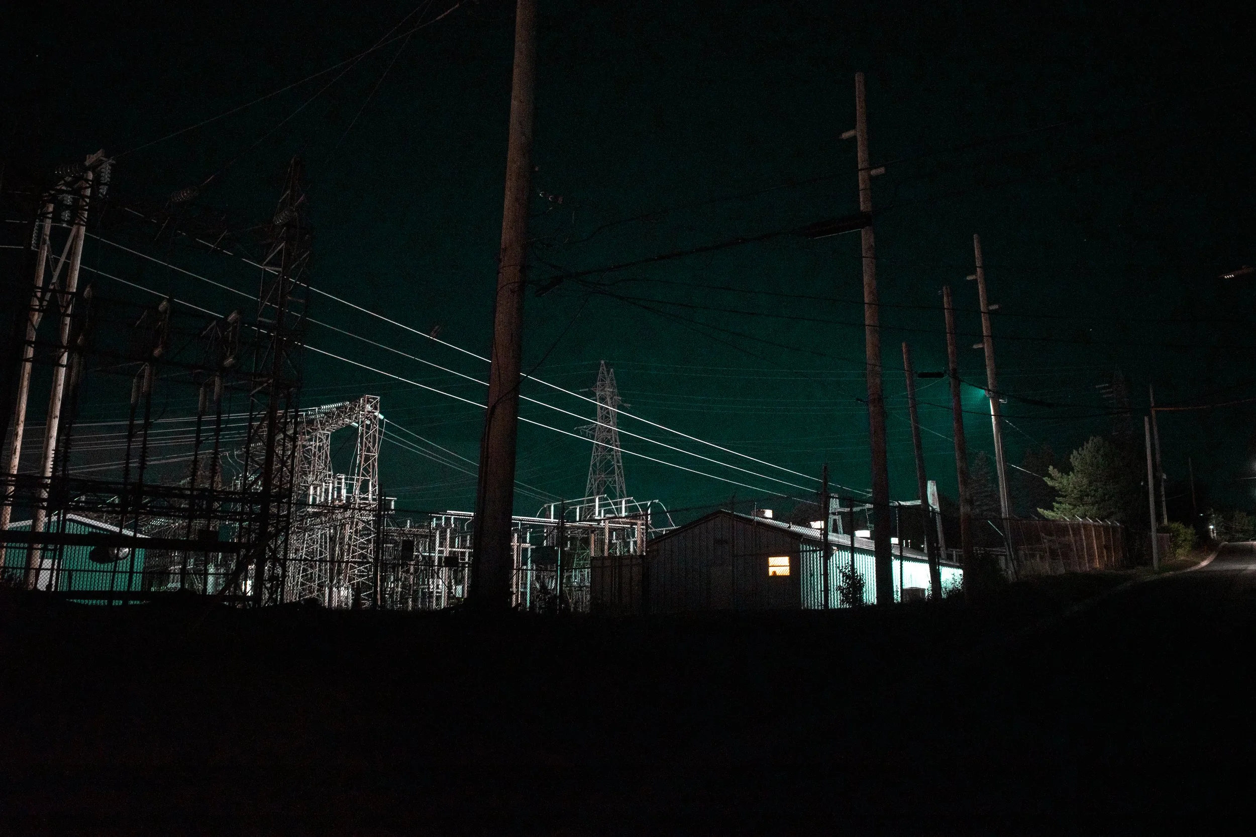 Night scene of electrical power substation with power lines and utility poles, illuminated by external light, on a dark road.