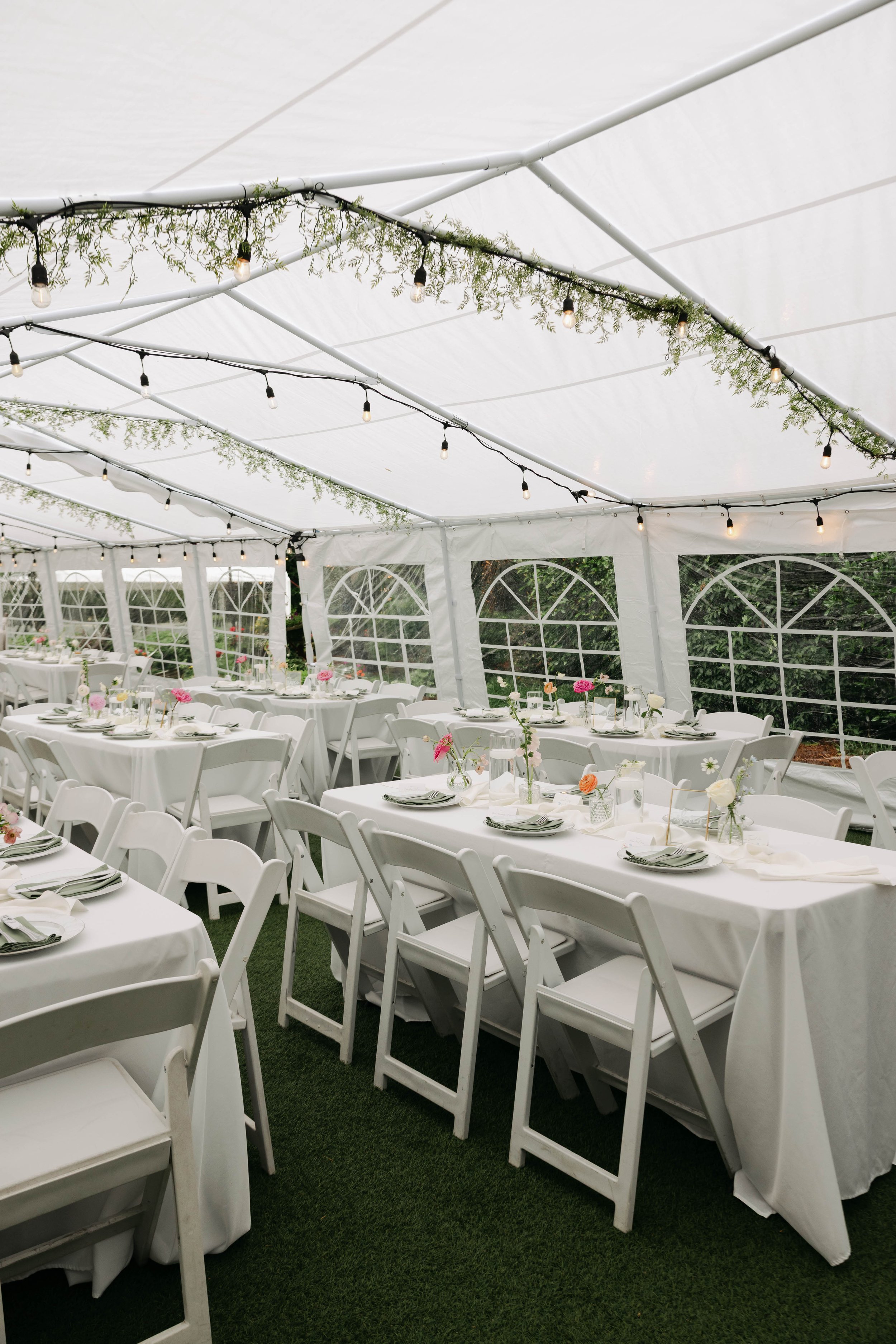 Inside a decorated white event tent with long tables covered in white tablecloths, set with plates, glasses, and floral centerpieces, and string lights hanging from the ceiling.