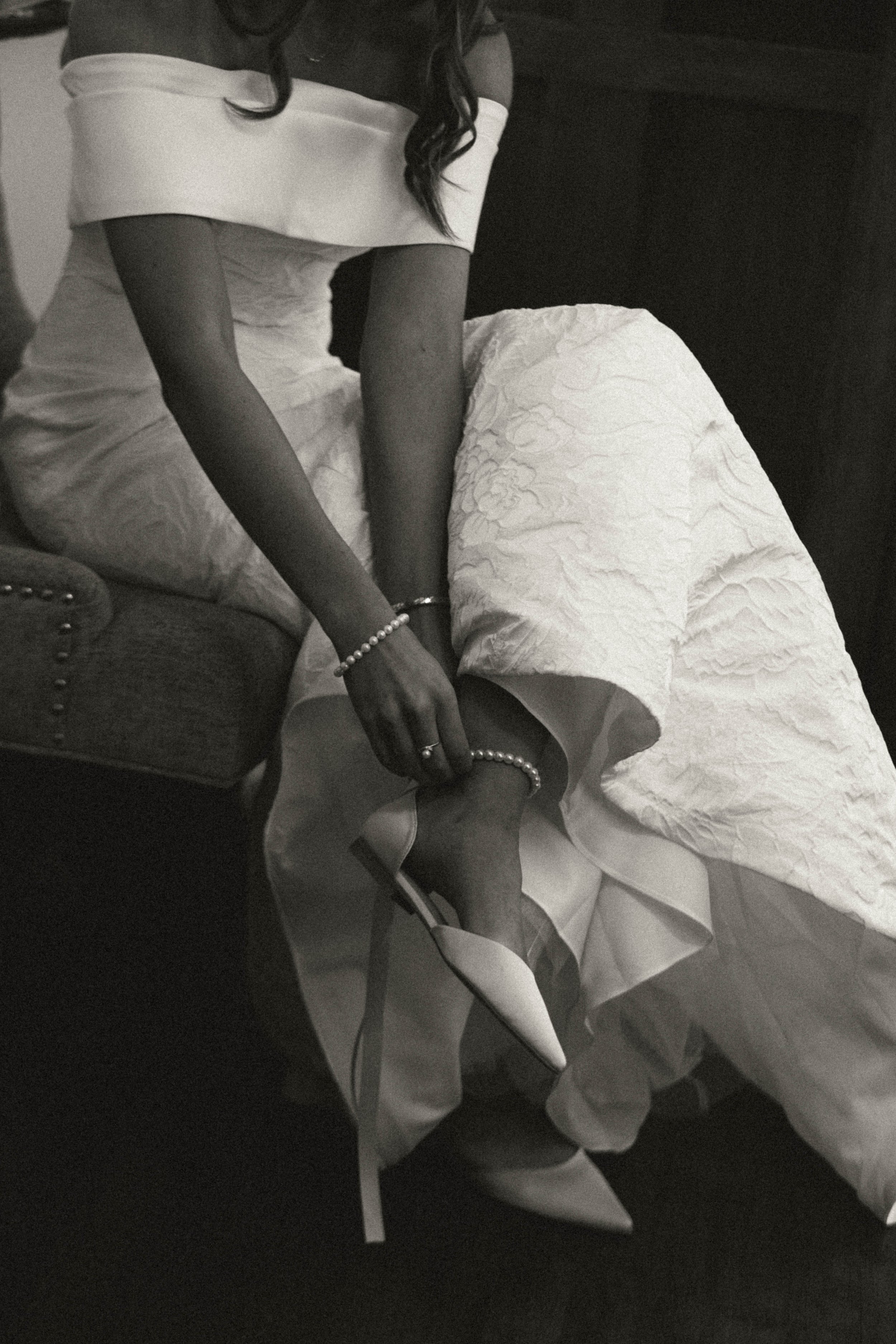 A woman in a black dress with an off-the-shoulder white overlay is sitting on a bed. She is wearing high-heeled shoes and holding a pearl necklace in her hand. The bed has a textured white cover, and the room is dimly lit.