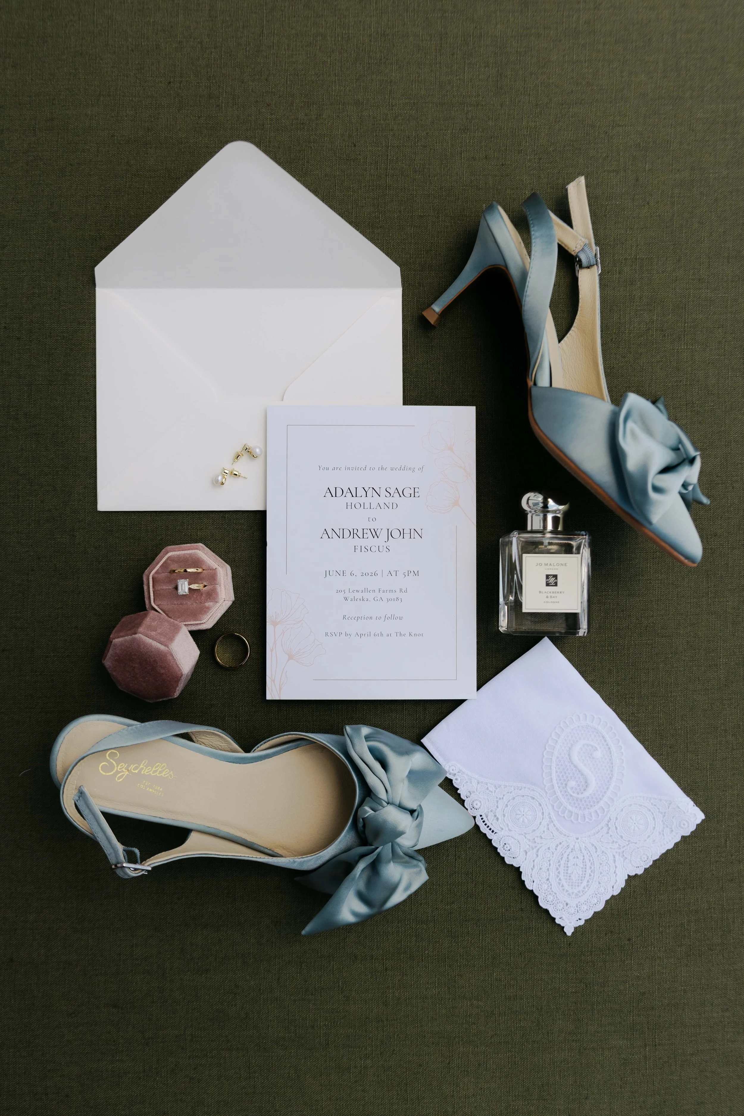 Wedding invitation, jewelry, blue satin high heels, perfume, and accessories on a dark green fabric background.