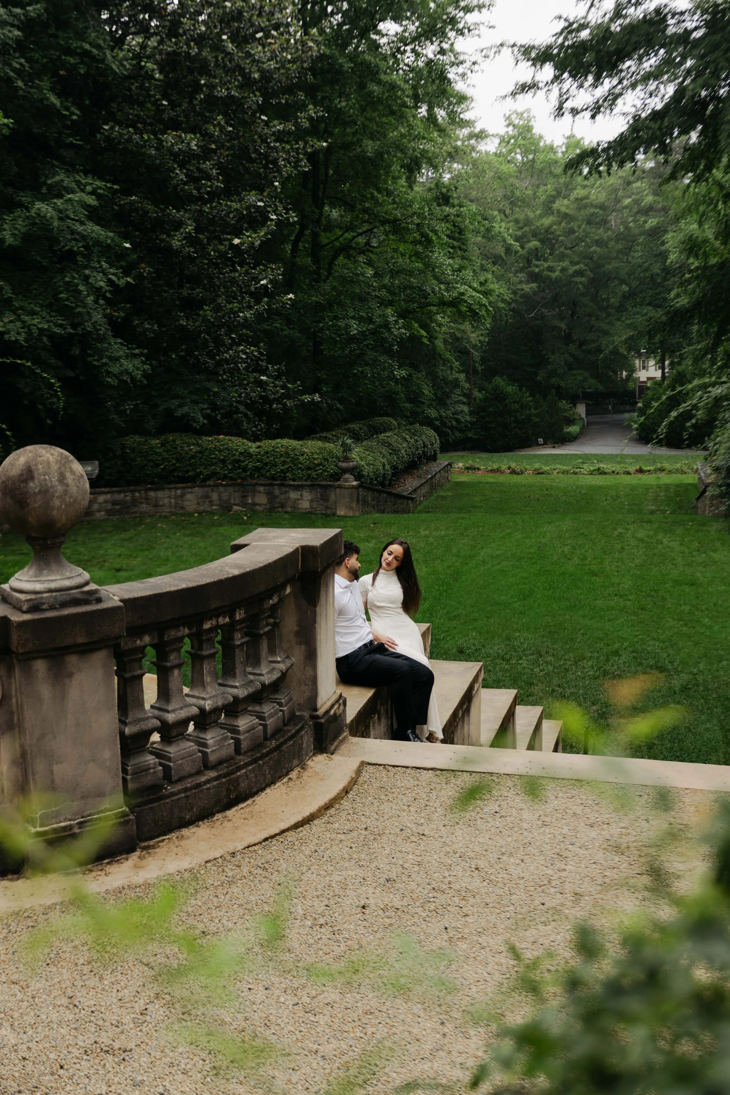 A couple sitting on stone steps by a stone balustrade in a lush green garden, with trees and a house in the background.