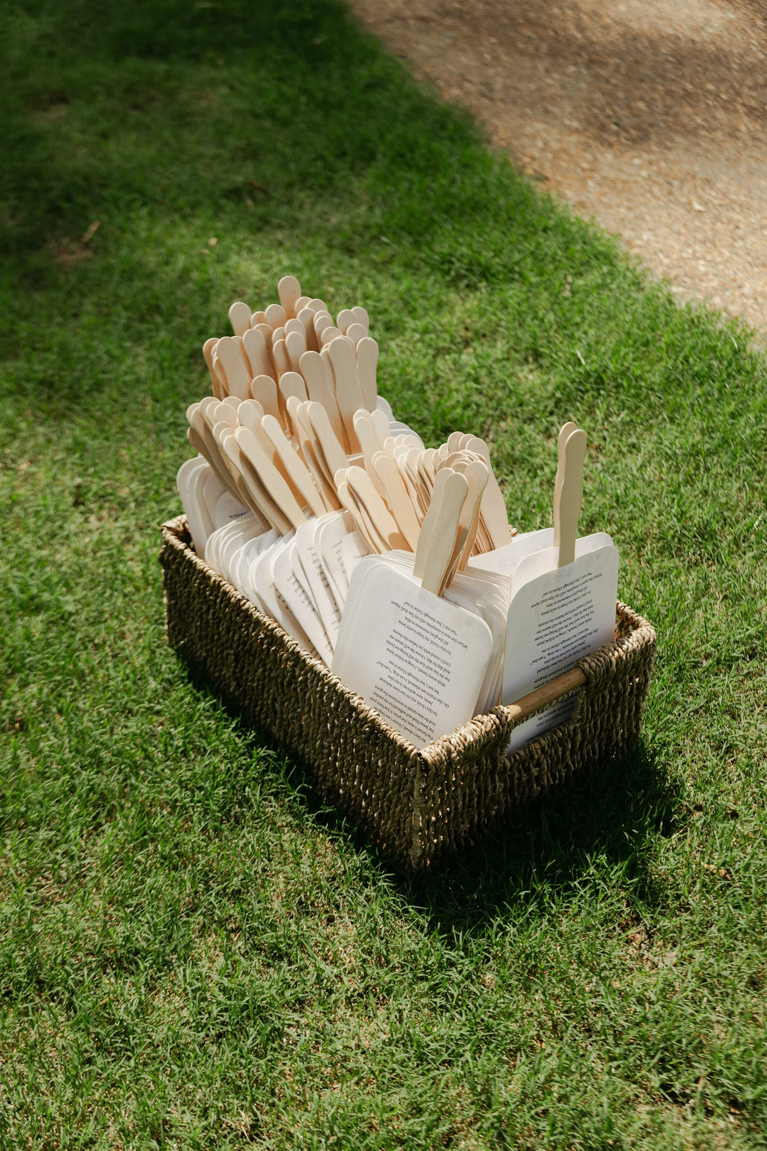 A basket filled with wooden popsicle sticks and paper tags rests on green grass next to a dirt path.