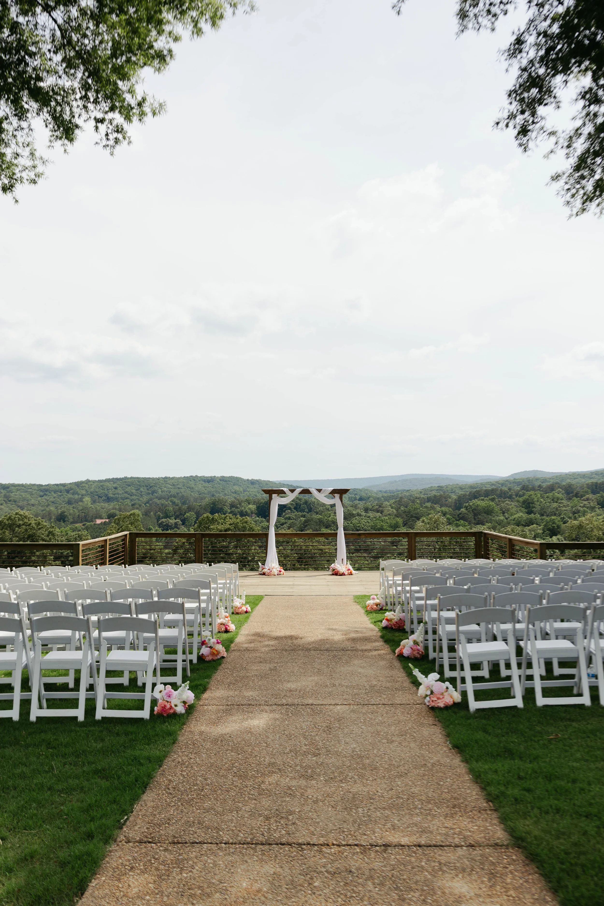 Outdoor wedding ceremony setup with white chairs arranged in rows on either side of a beige aisle, decorated with pink and white flowers, leading to a wooden wedding arch with white fabric, set against a backdrop of green hills and a cloudy sky.
