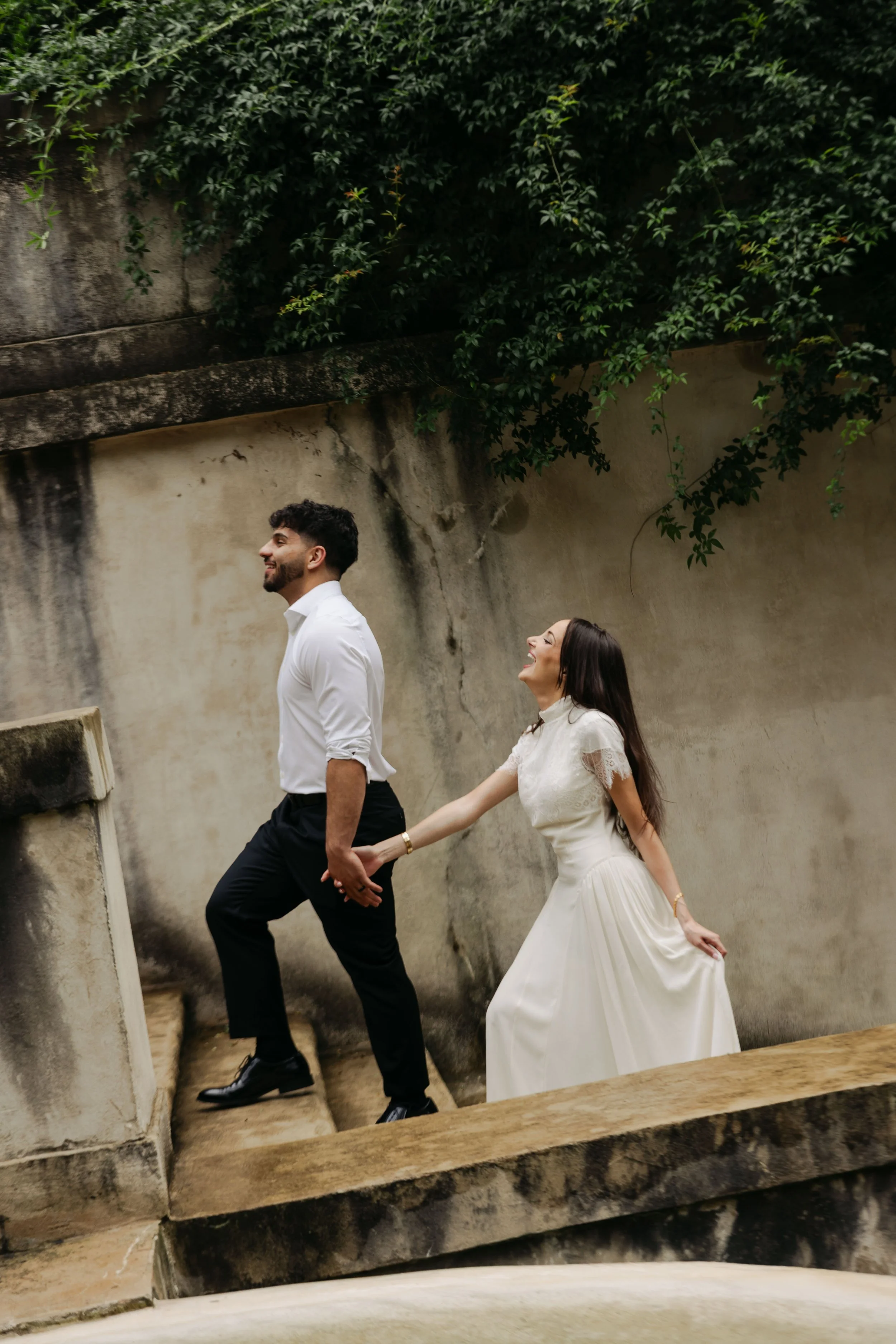A man and woman enjoying a walk outdoors, holding hands and smiling, with a rustic stone wall and lush green foliage in the background.