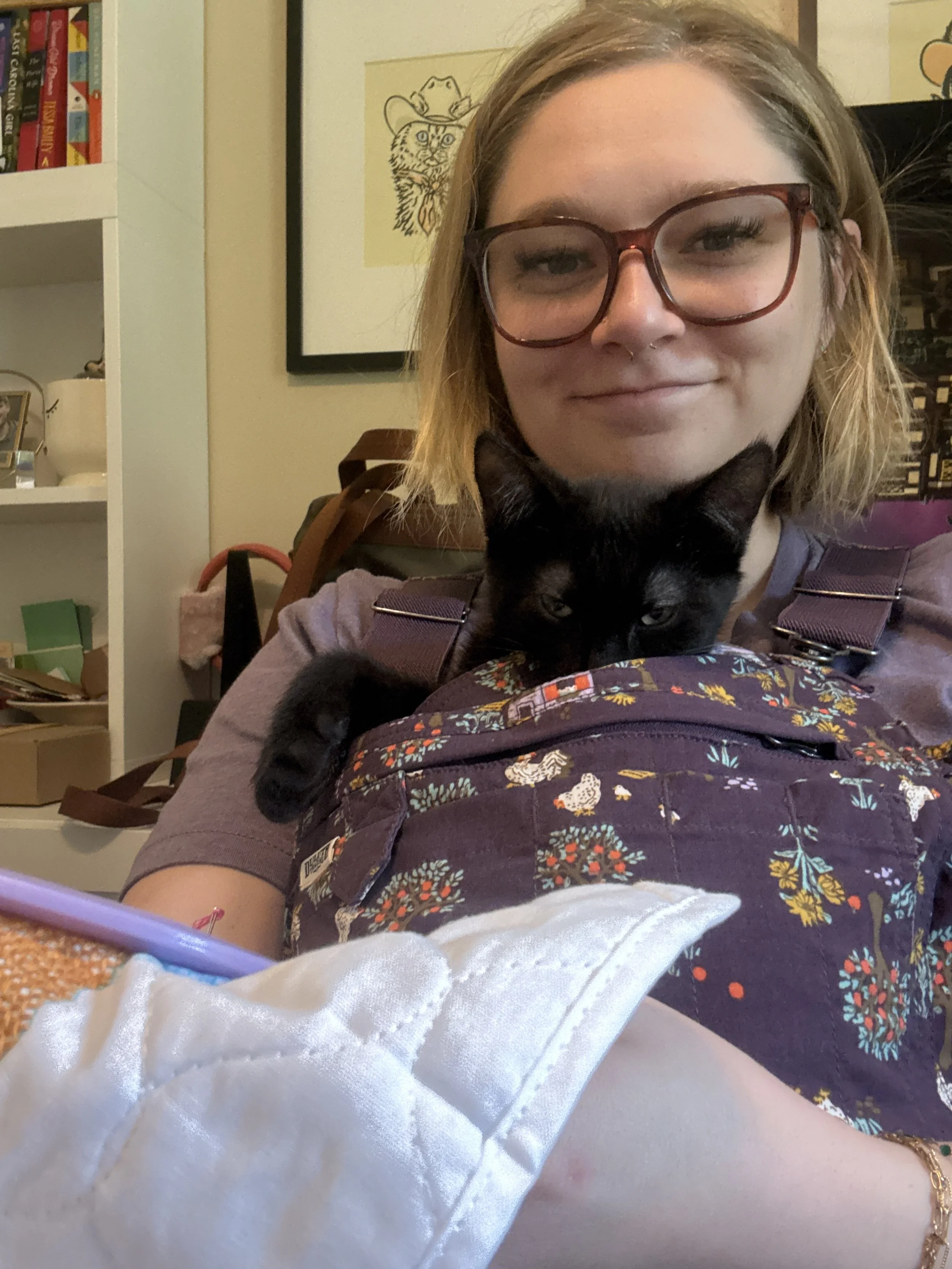 A woman with glasses and a septum piercing holding a black cat in a purple floral backpack in a room with books and artwork on the walls.