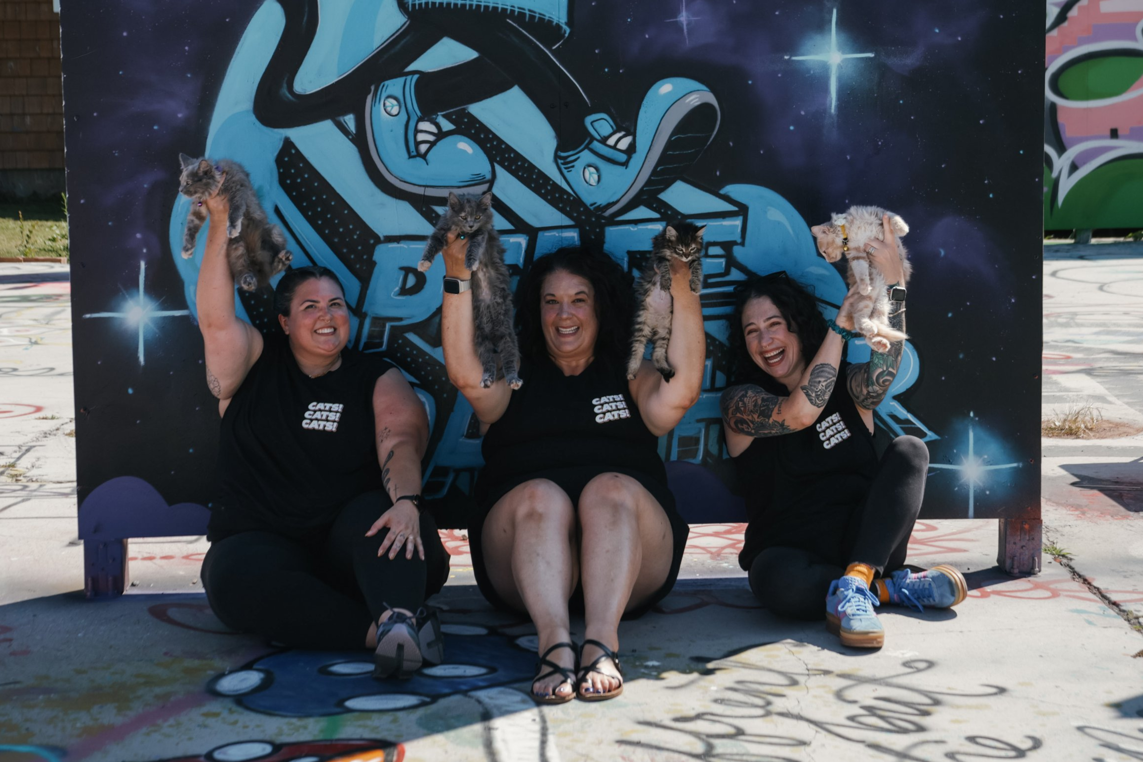 Three women sitting on the ground in front of a space-themed mural, each holding three cats, smiling and raising their arms. The women are wearing black shirts with white text, and the cats are of various colors and patterns.