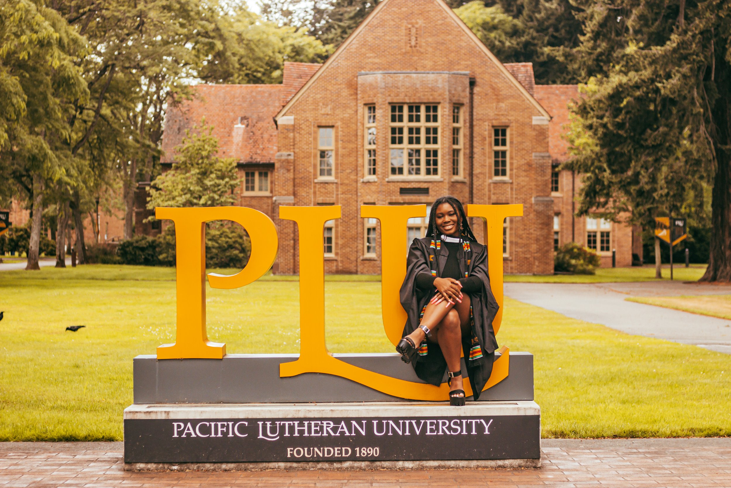 A Black student who is graduating with their masters degree sitting on a PLU sign (standing for Pacific Lutheran University) with their legs crossed.