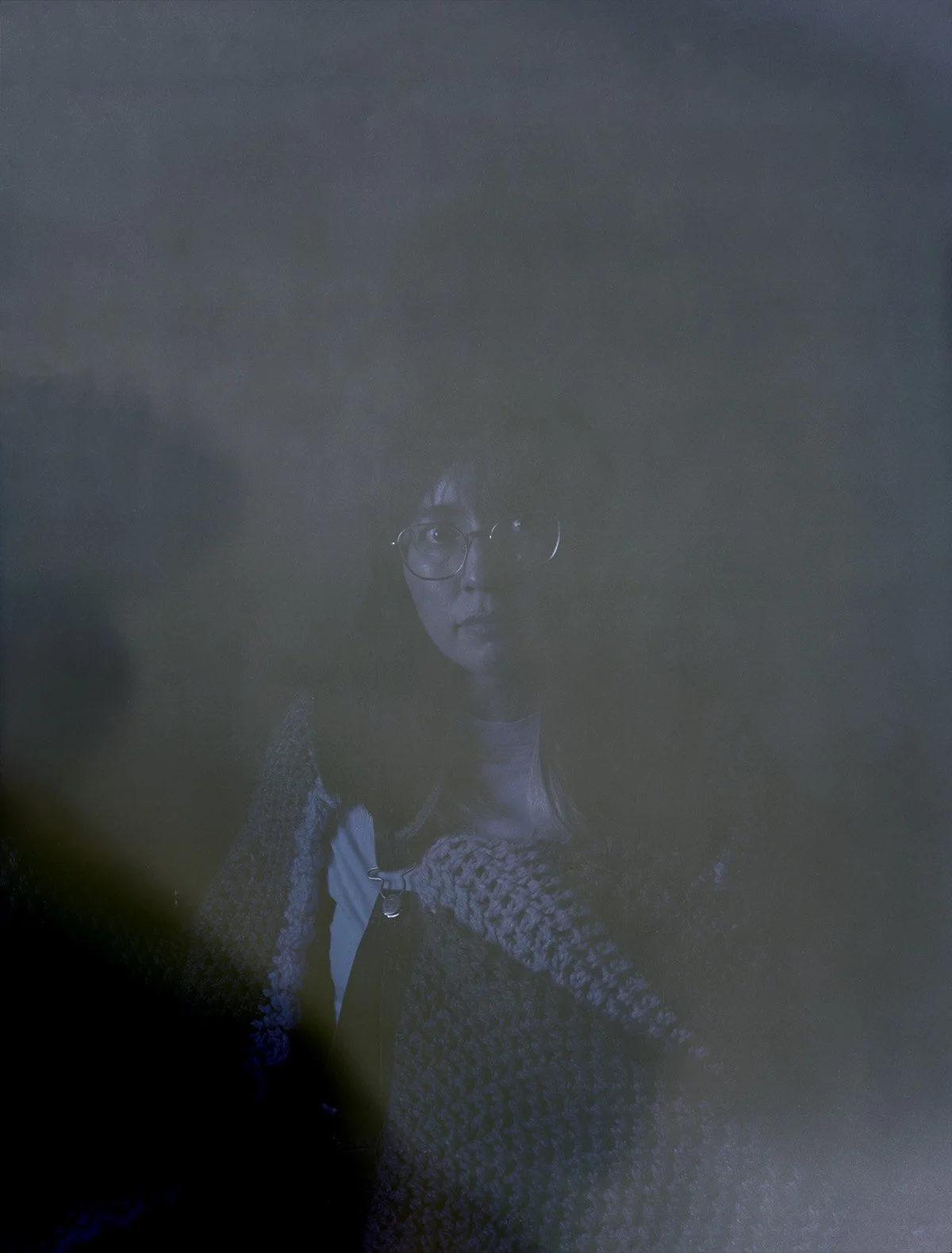 A woman wearing glasses and a patterned jacket, with short dark hair, illuminated subtly in a dark environment.