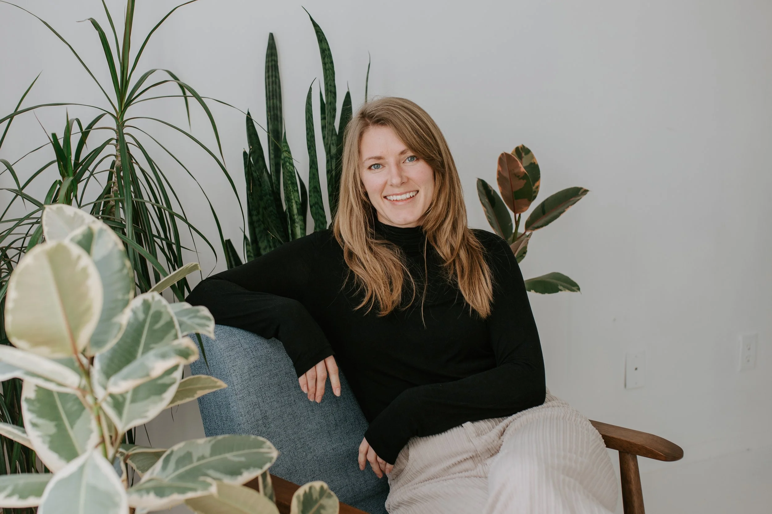 A woman with long blonde hair and blue eyes sitting in a room with a gray sofa, in front of a plain light gray wall, surrounded by large green houseplants, smiling at the camera.